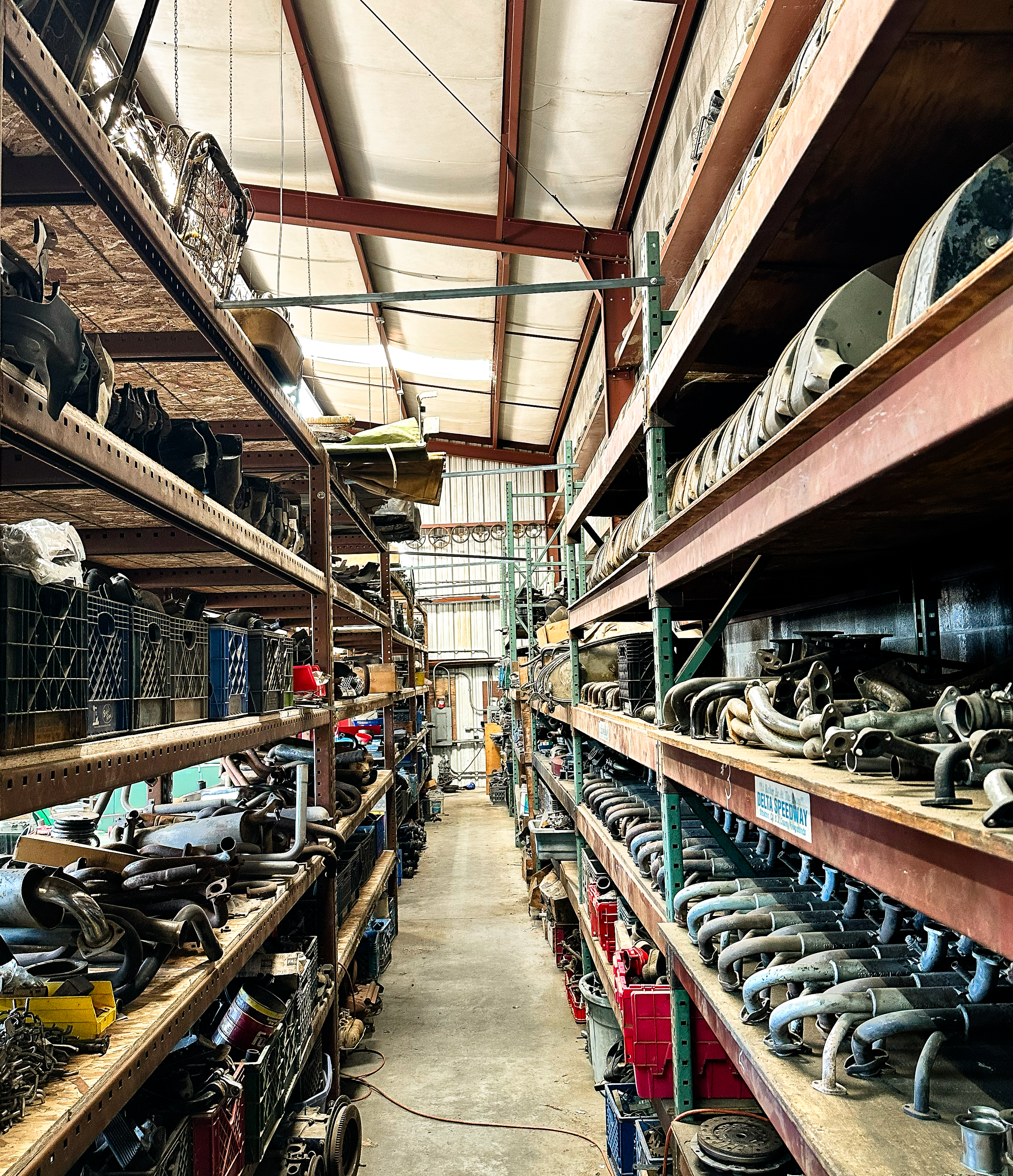 Aisle in a warehouse or parts store with shelves stocked with vehicle auto parts, metal pipes, and tools.
