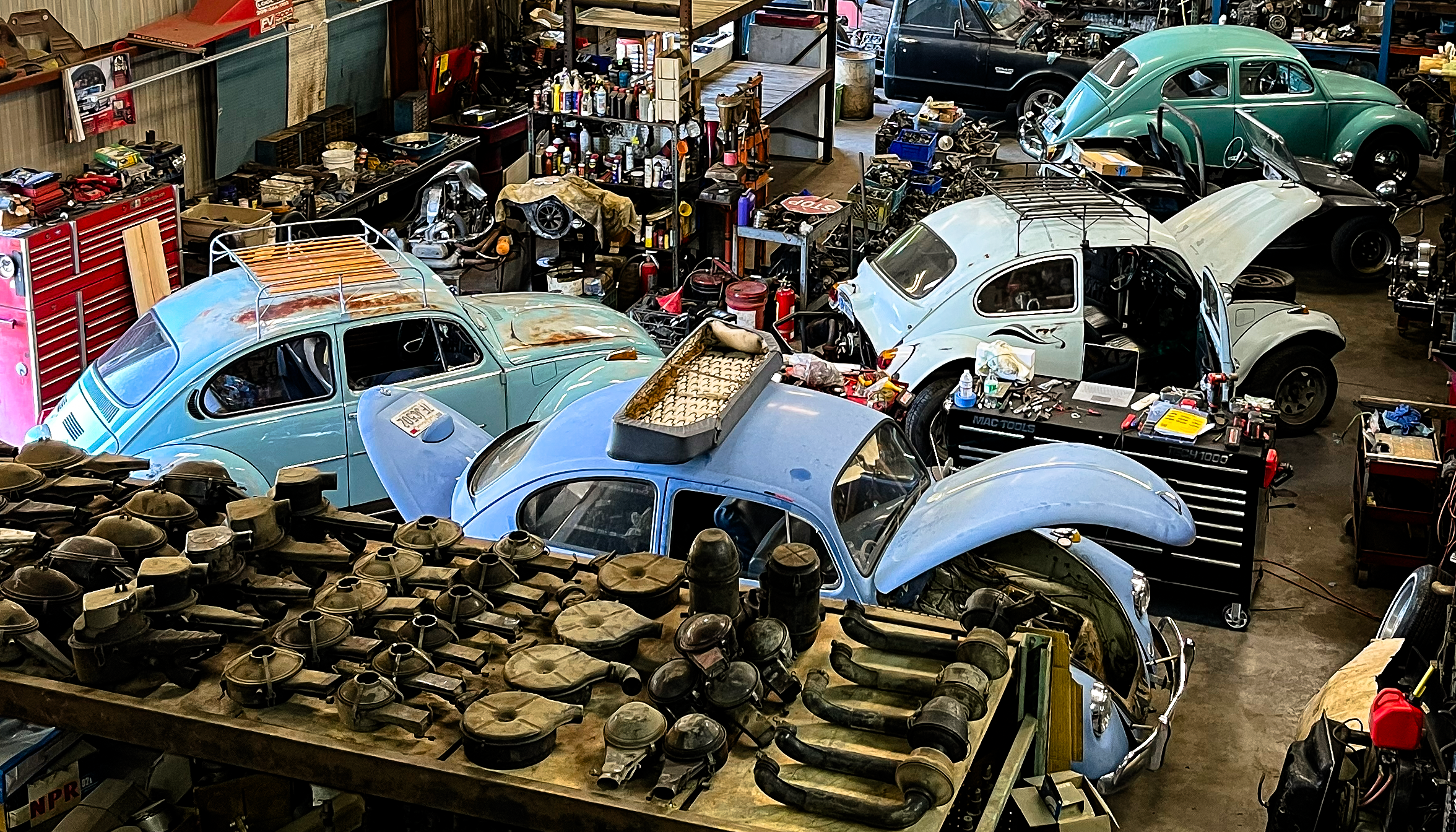 A garage filled with vintage cars in various stages of repair, with numerous tools and car parts scattered around.