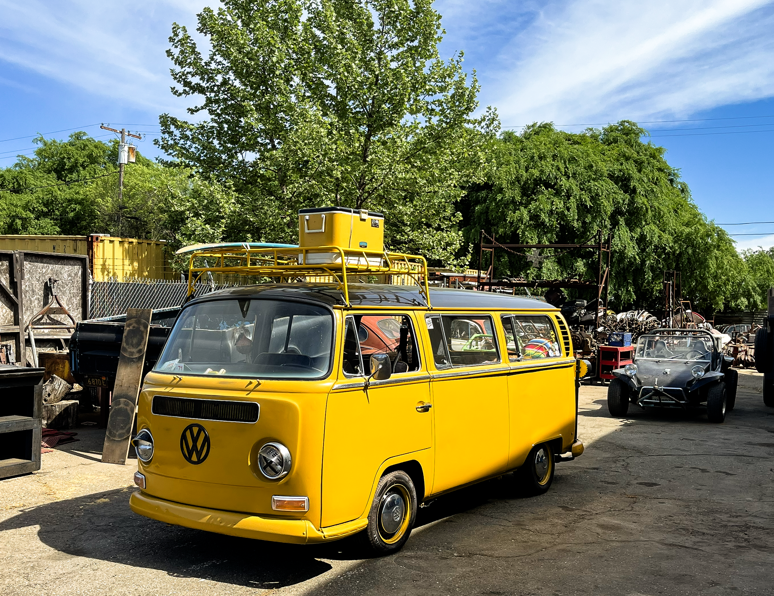 A vibrant yellow vintage Volkswagen van parked in an outdoor lot surrounded by trees and various other vehicles and machinery under a clear blue sky.
