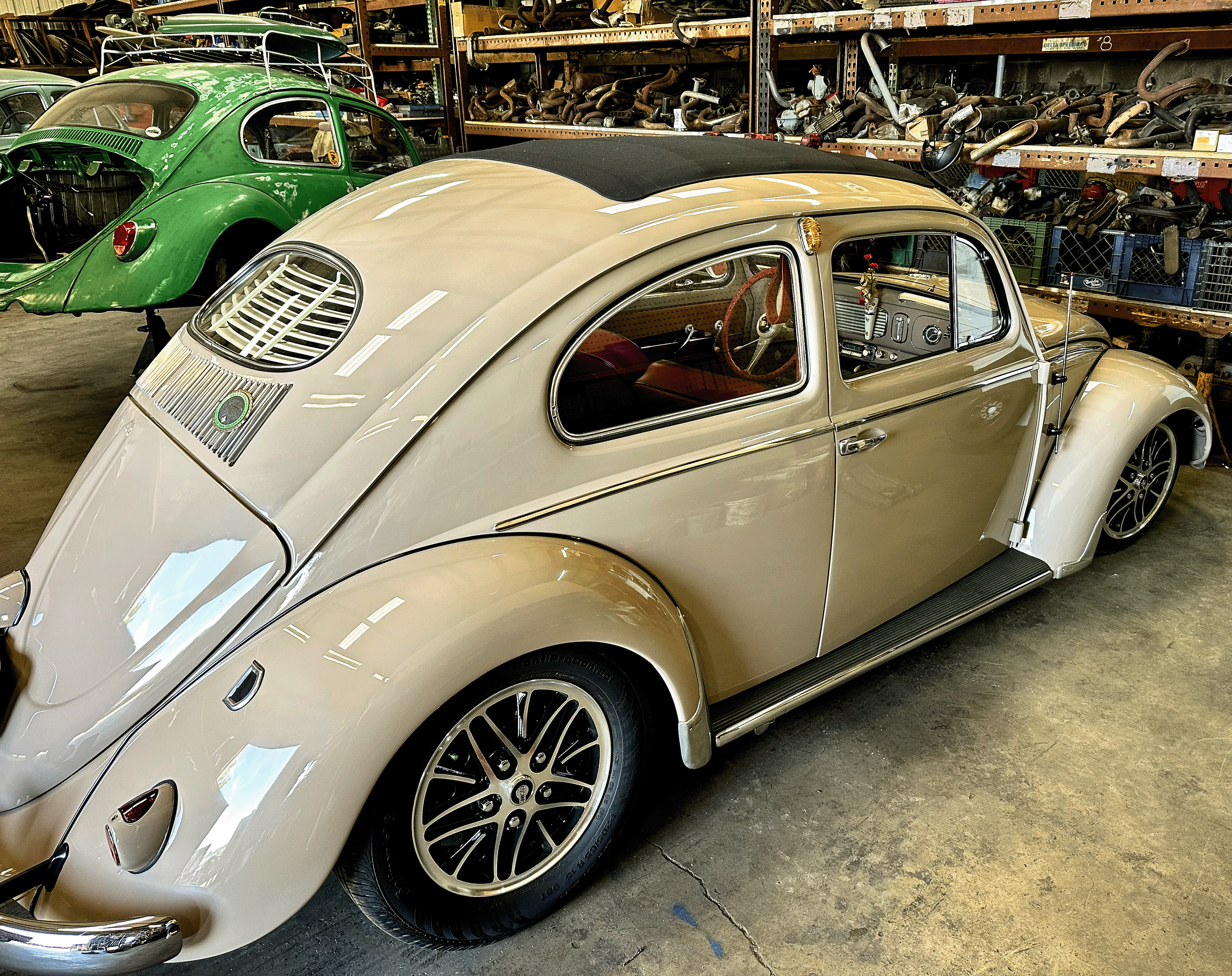 A vintage cream-colored Volkswagen Beetle with a black roof, parked inside a storage warehouse filled with car parts and other vehicles. The Beetle has distinctive round headlights, a shiny exterior, and custom wheels.