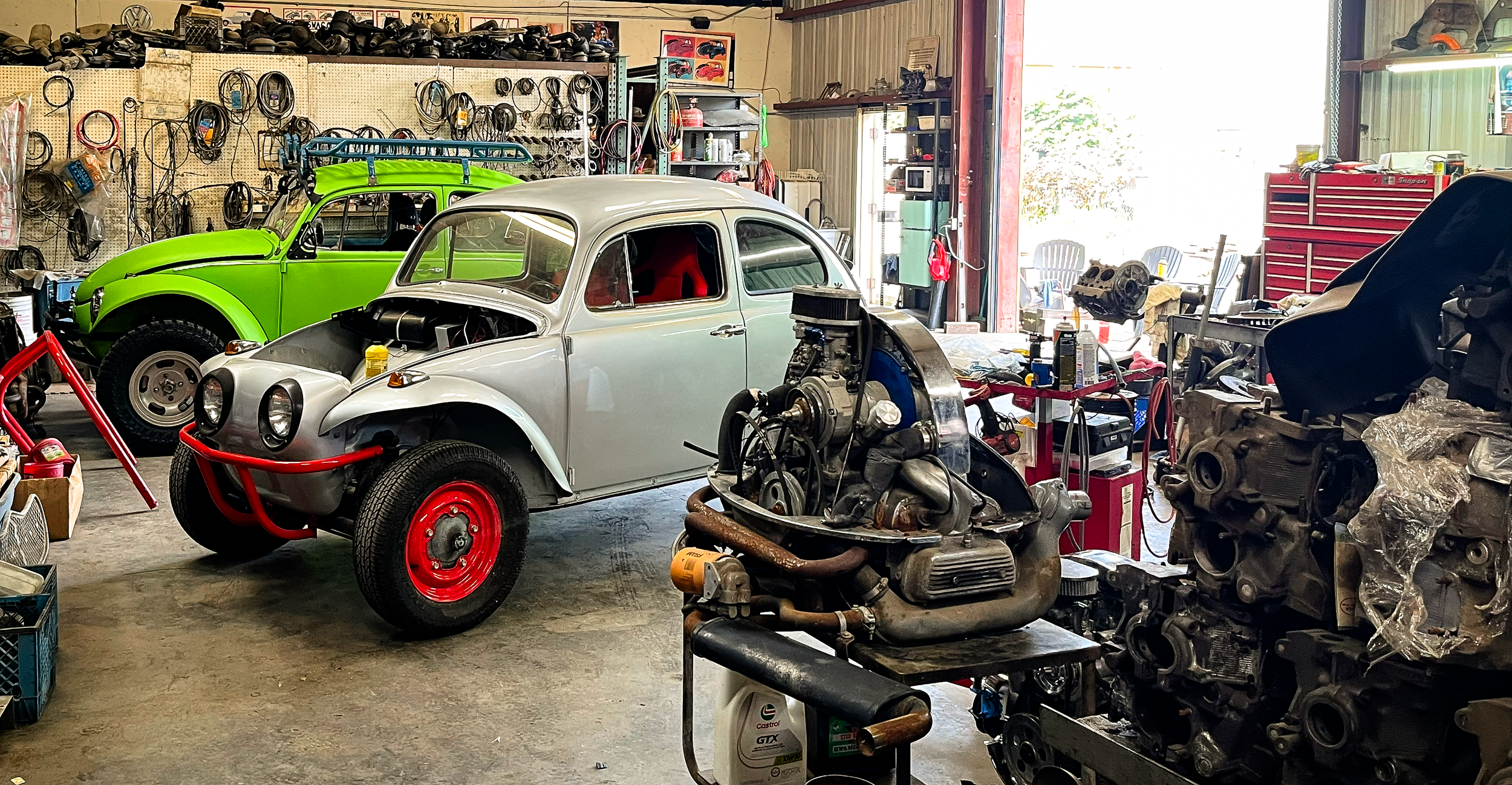 A garage workshop with two partially assembled classic Volkswagen Beetles, one painted bright green and the other in primer with red wheels. The space is cluttered with car parts, tools, and equipment, with a pegboard wall filled with various cables 