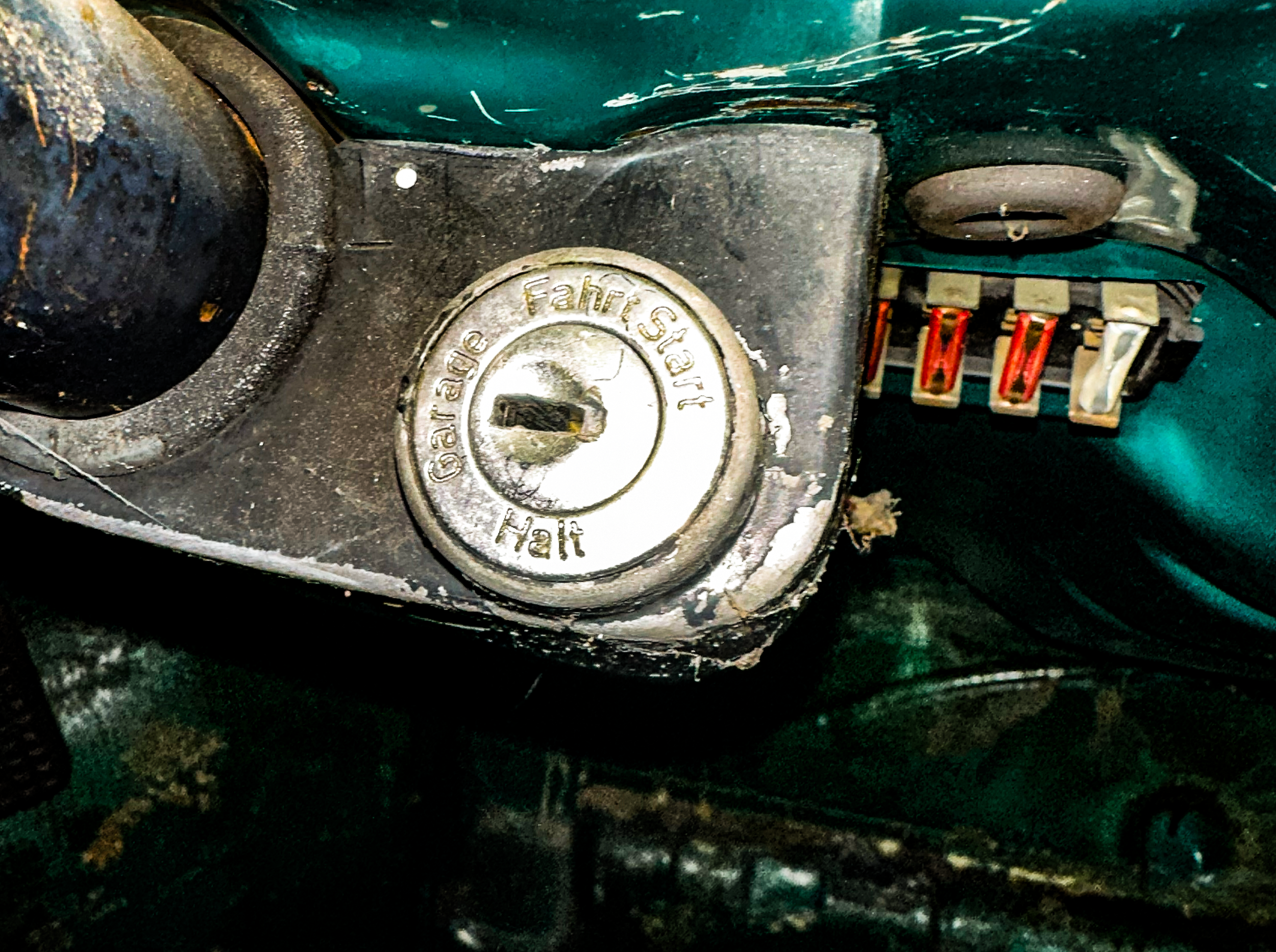 Close-up of an old, weathered lock and keyhole with the words 'Fairlock' engraved, and a small part of a rusty, antiquated-locking mechanism with wires nearby, on a dark, worn surface.
