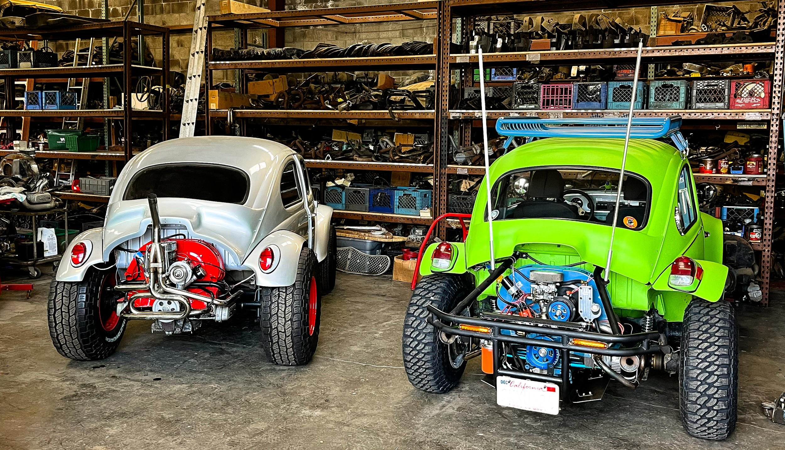 A garage with two dune buggies, one silver and one bright green, parked in front of shelves filled with car parts, tools, and plastic storage bins.