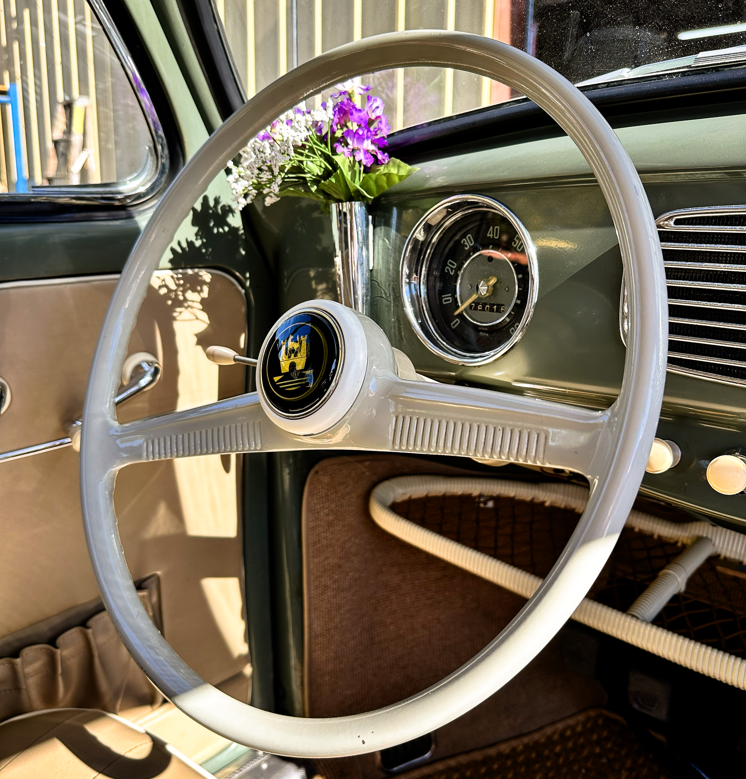 Inside a vintage car, the dashboard displays a speedometer, and a white steering wheel with the Ford logo. A small vase with purple and white flowers decorates the dashboard. The car interior includes a beige seat and a view of a wooden fence outside