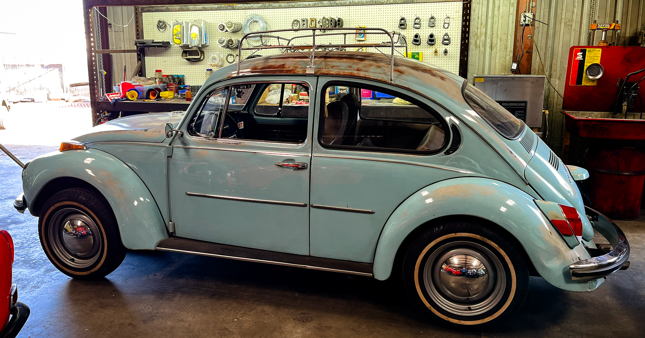 A vintage light blue Volkswagen Beetle with a rusty roof, parked inside a garage or workshop, with shelves and tools in the background.