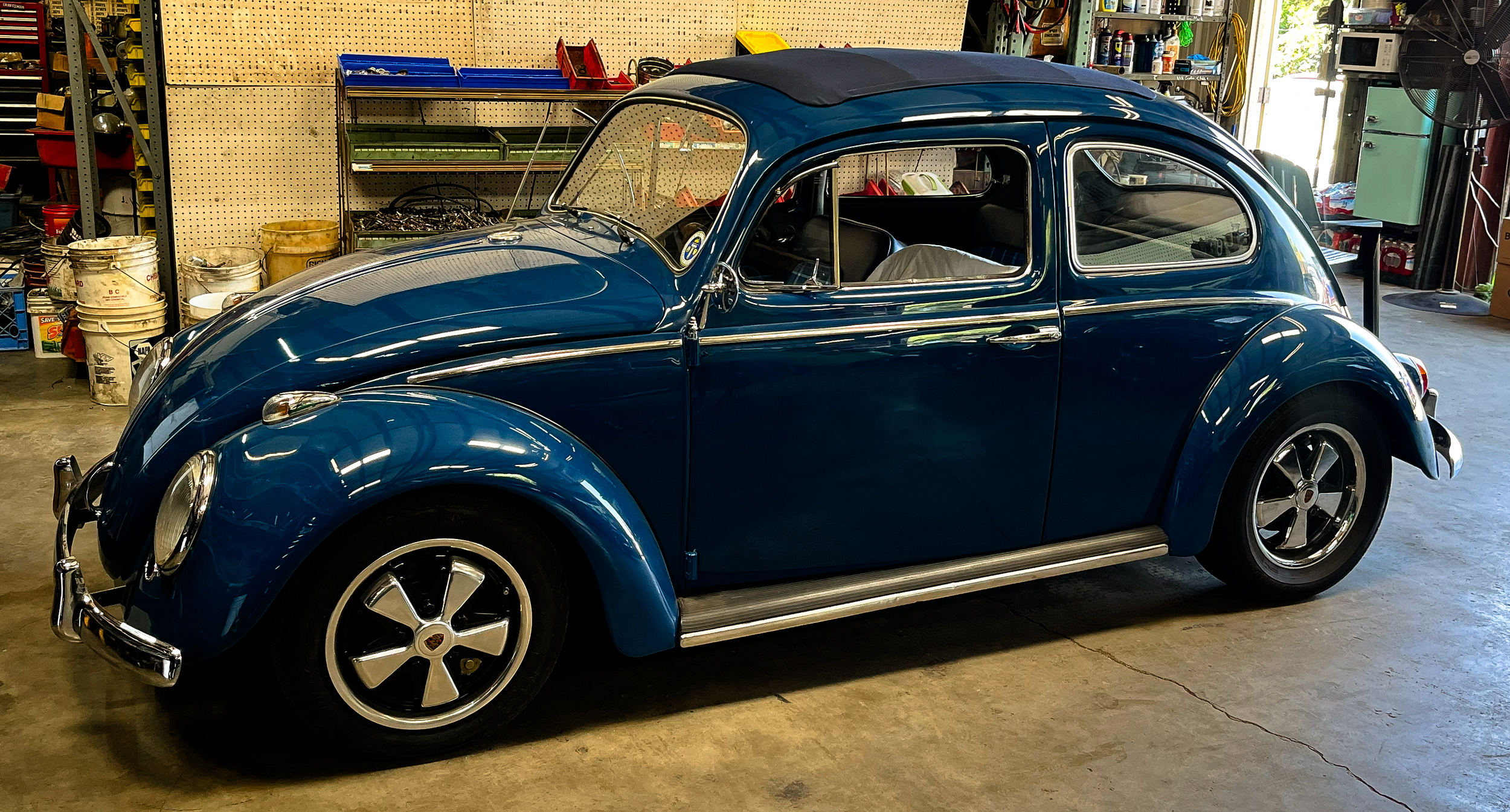 A blue vintage Volkswagen Beetle car parked inside a garage with tools and supplies stored on shelves and containers.