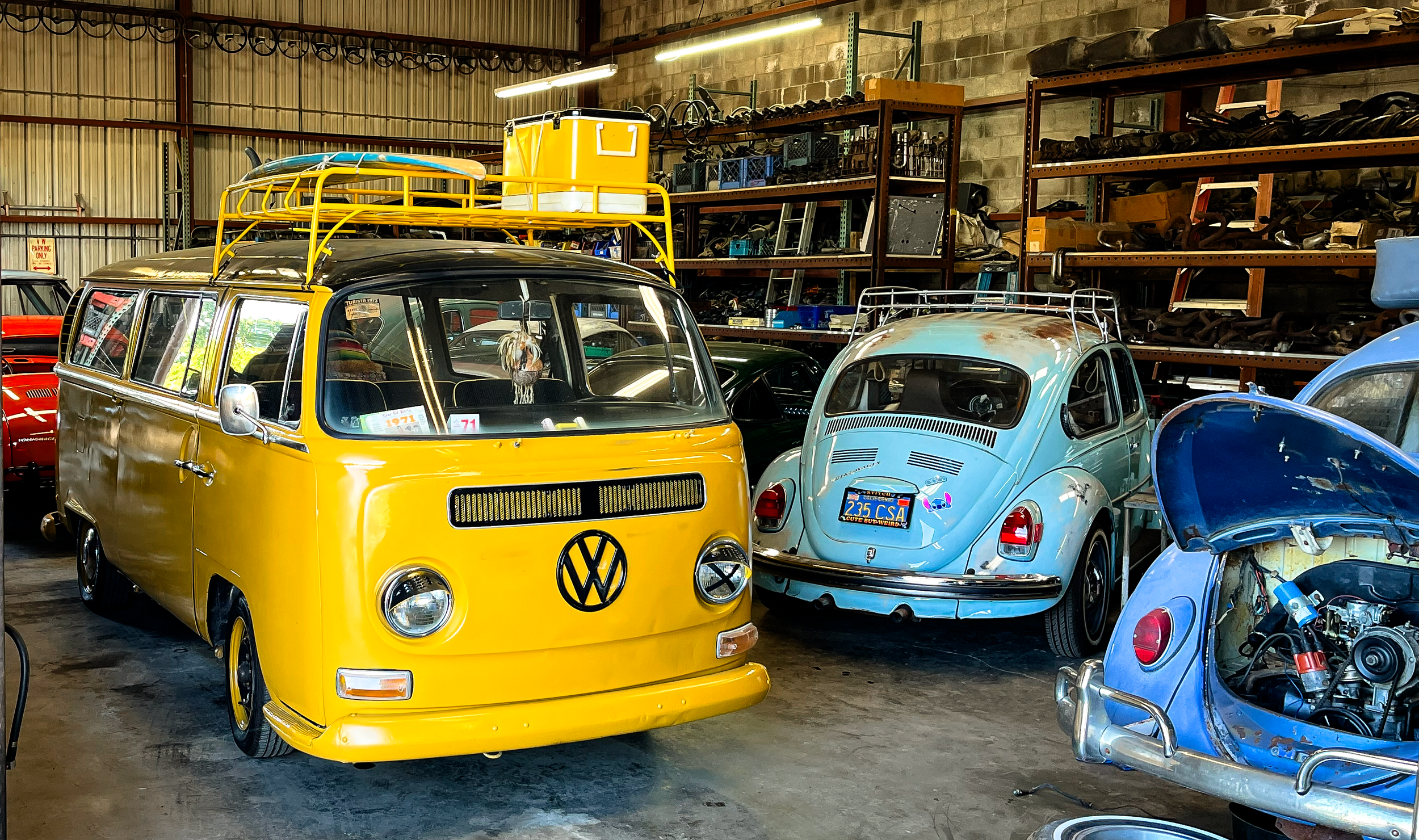 Interior of a storage warehouse with vintage Volkswagen vehicles, including a bright yellow van with a roof rack, a light blue Beetle with a California license plate, and other classic cars stored on shelves and on the floor.