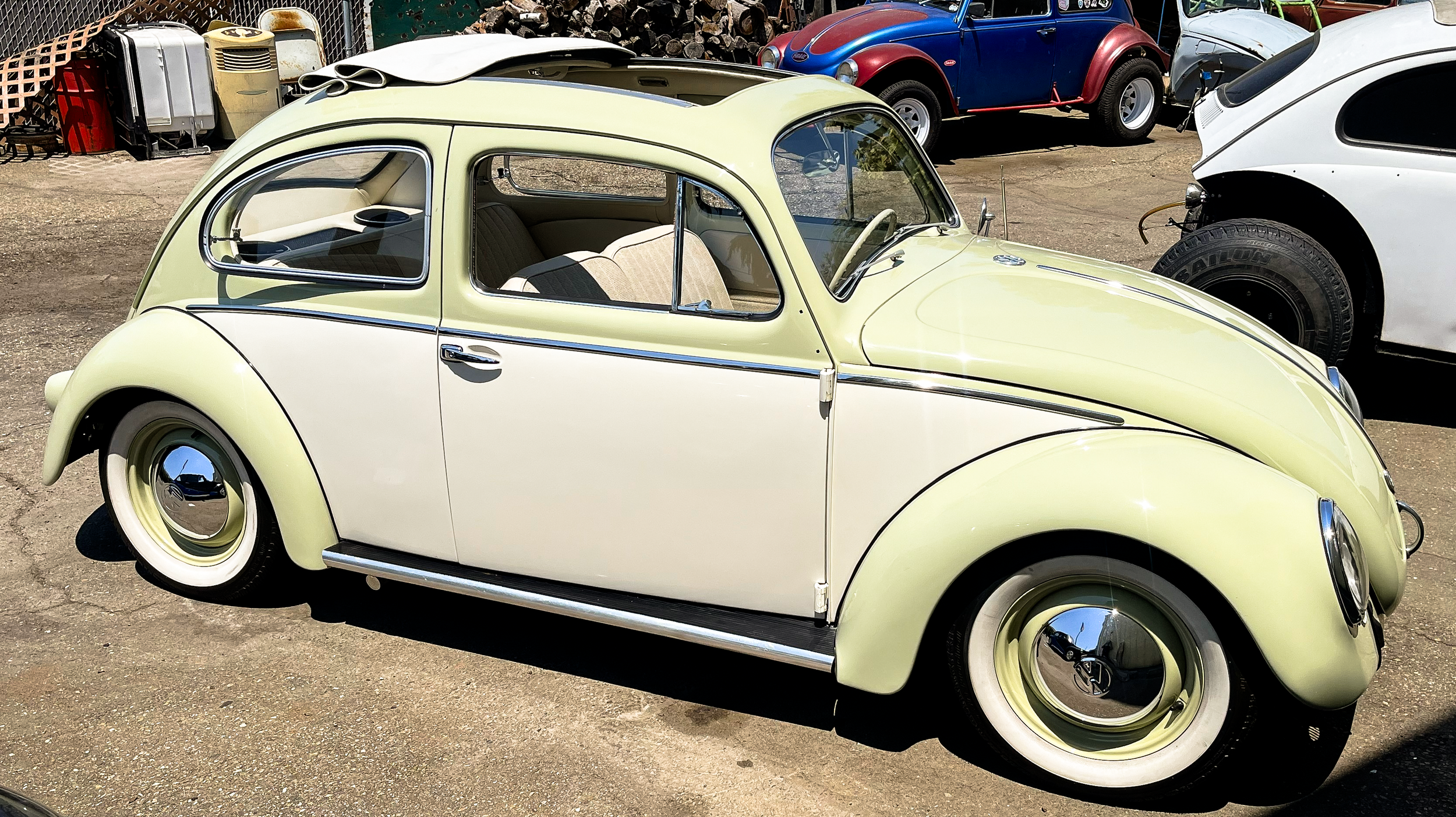 A vintage pale yellow and white Volkswagen Beetle parked outdoors among other classic cars, with a stack of logs and various equipment in the background.