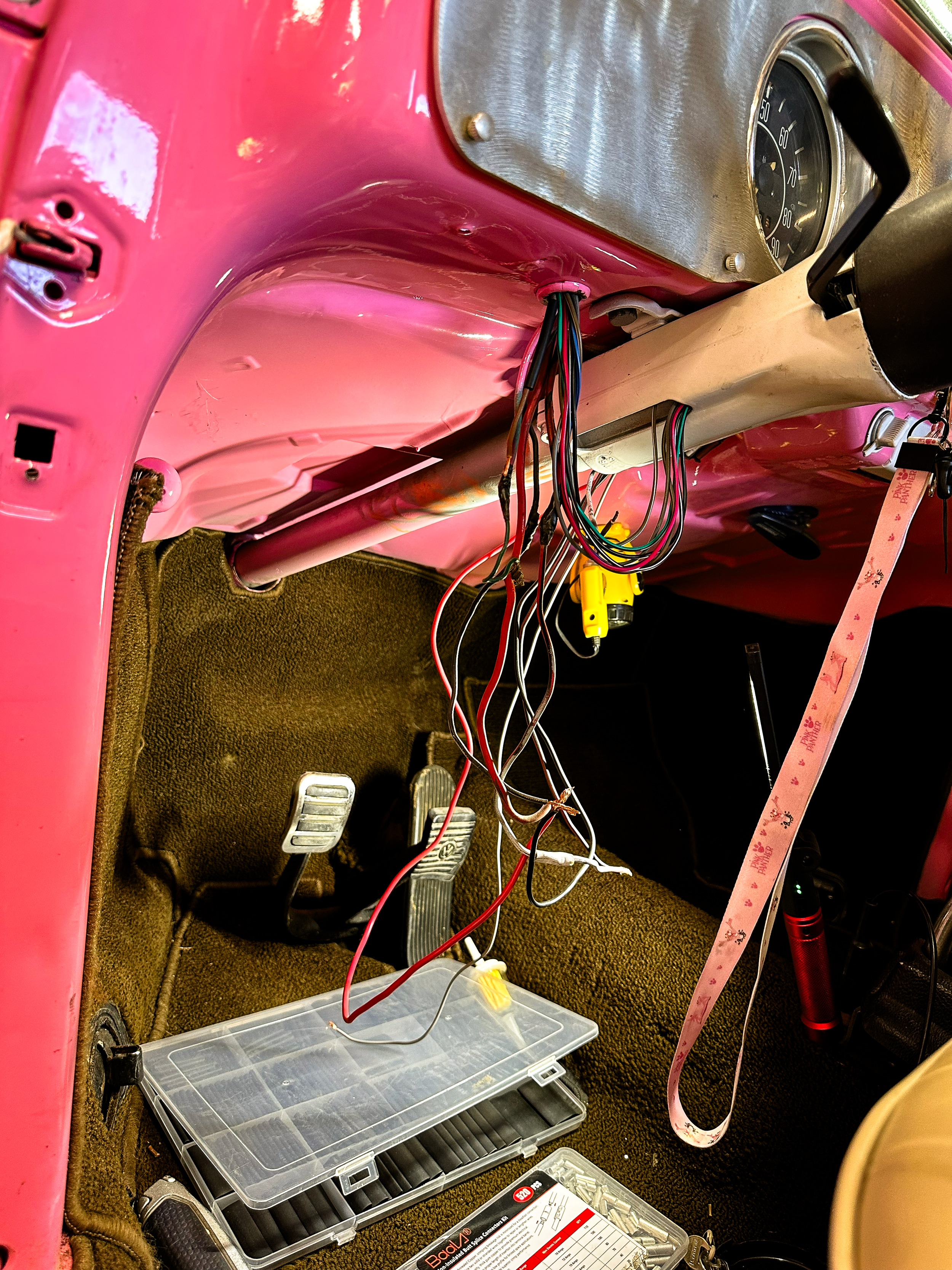 The image shows the interior of a vintage pink car with the dashboard partially disassembled. There are multiple loose wires hanging and tools, including a yellow screwdriver and a clear plastic toolbox containing various screws and parts, placed on 