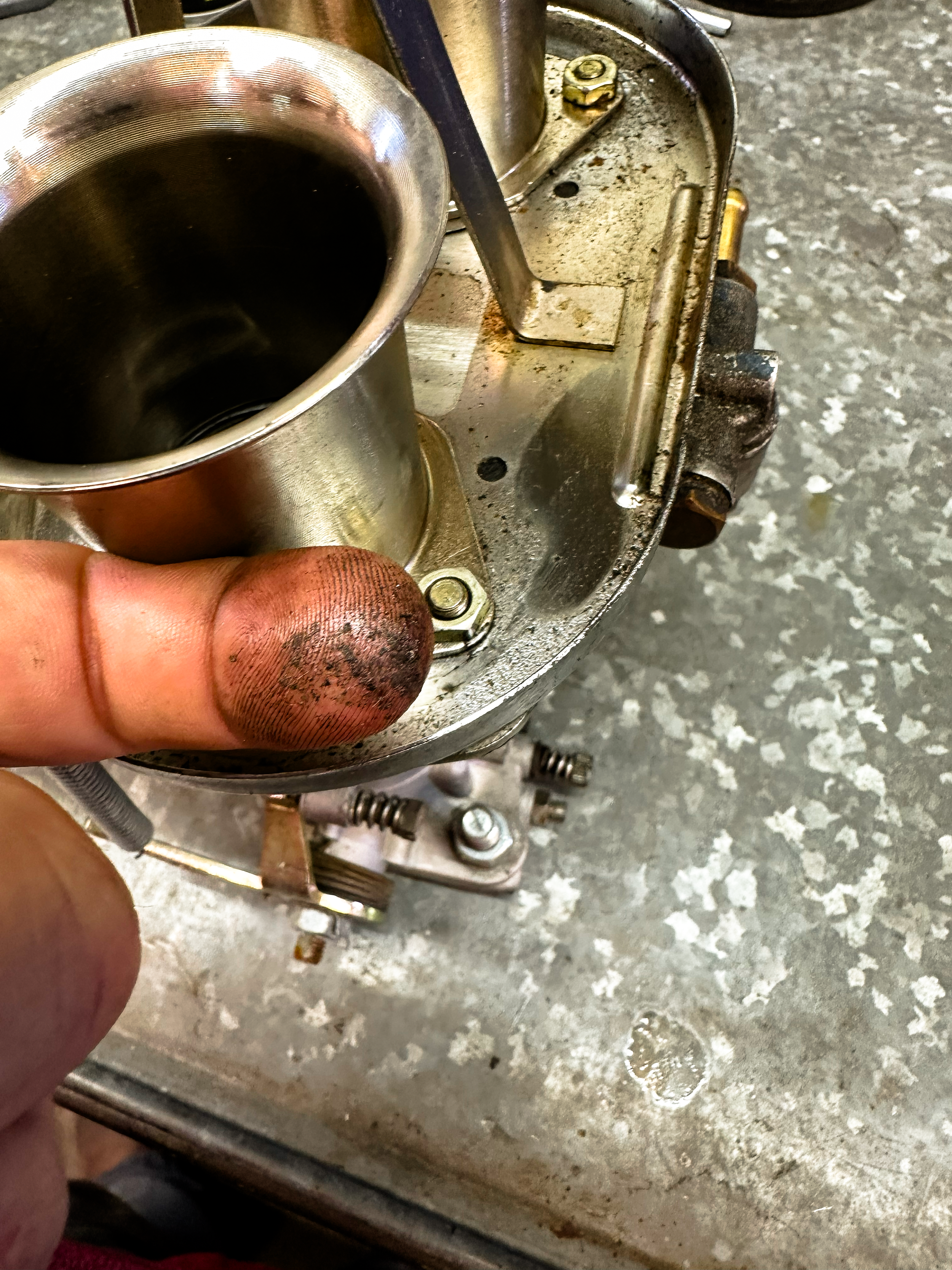 Close-up of a person's finger holding a metal component, part of a mechanical or engine assembly, with visible oil stains and a rusty bolt, on a work surface with scattered screws and a worn metallic surface.