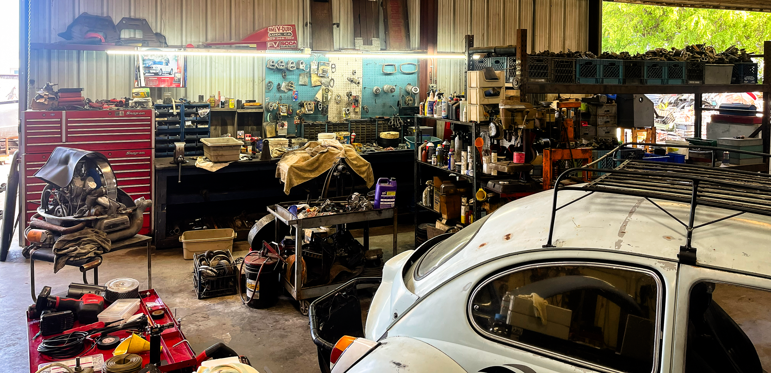 A cluttered garage workshop with tools, car parts, and equipment. A vintage white car and a red tool chest are visible, with shelves and a pegboard full of various items.