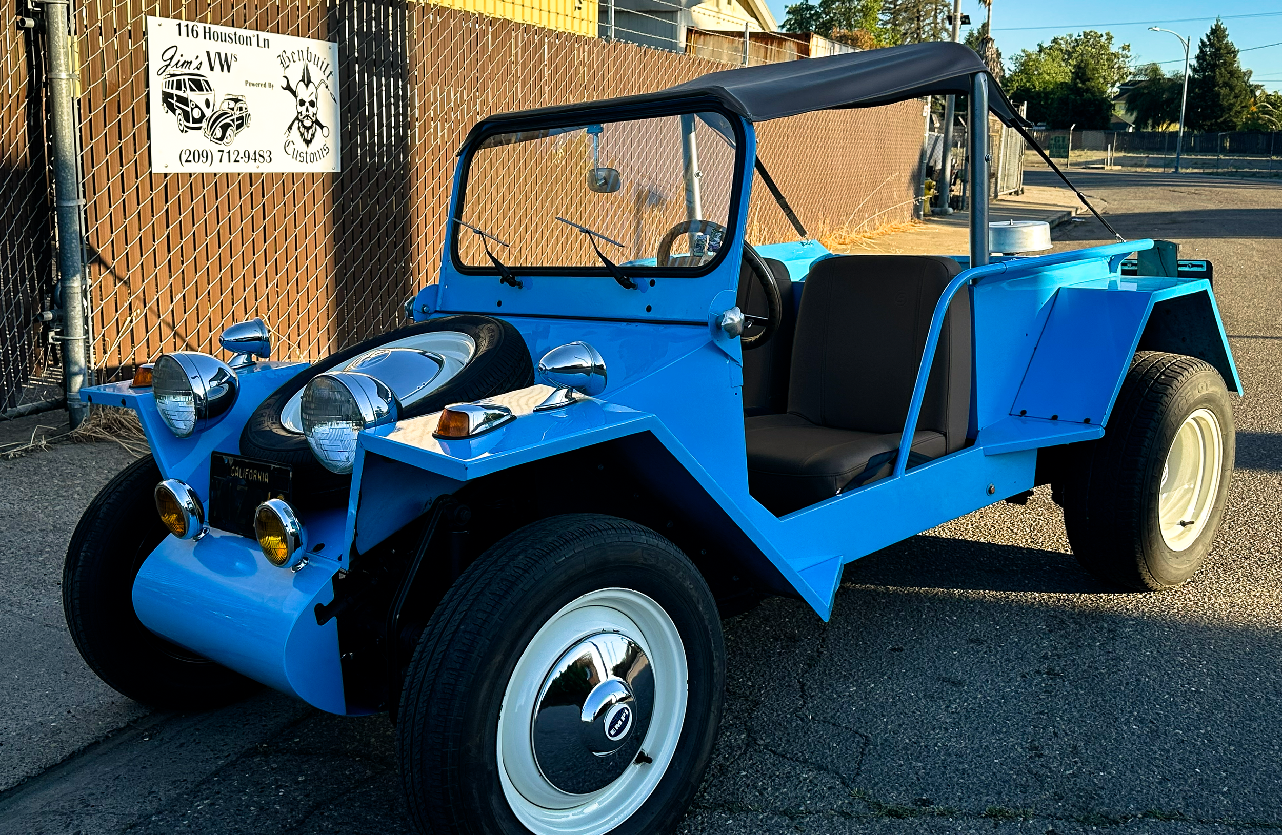 A bright blue vintage golf cart parked outdoors near a chain-link fence and a sign that reads "116 Houston Ln Jim's VW Customs," with a phone number. The golf cart has a black canopy, four large white wheels, and classic round headlights.