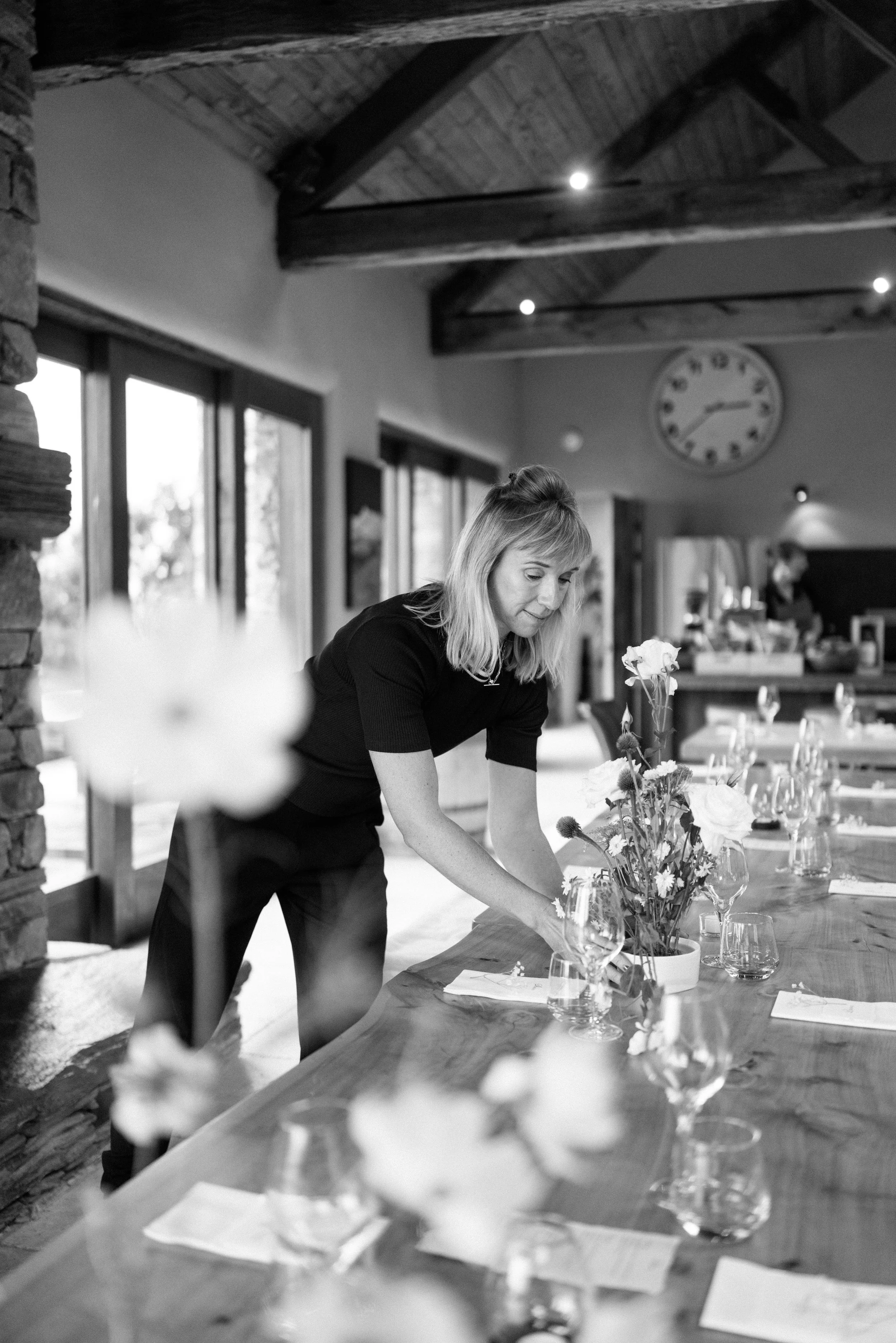 Anna arranging flowers in a vase on a long wooden table in a rustic room with wooden beams and large windows, with a clock on the wall in the background.
