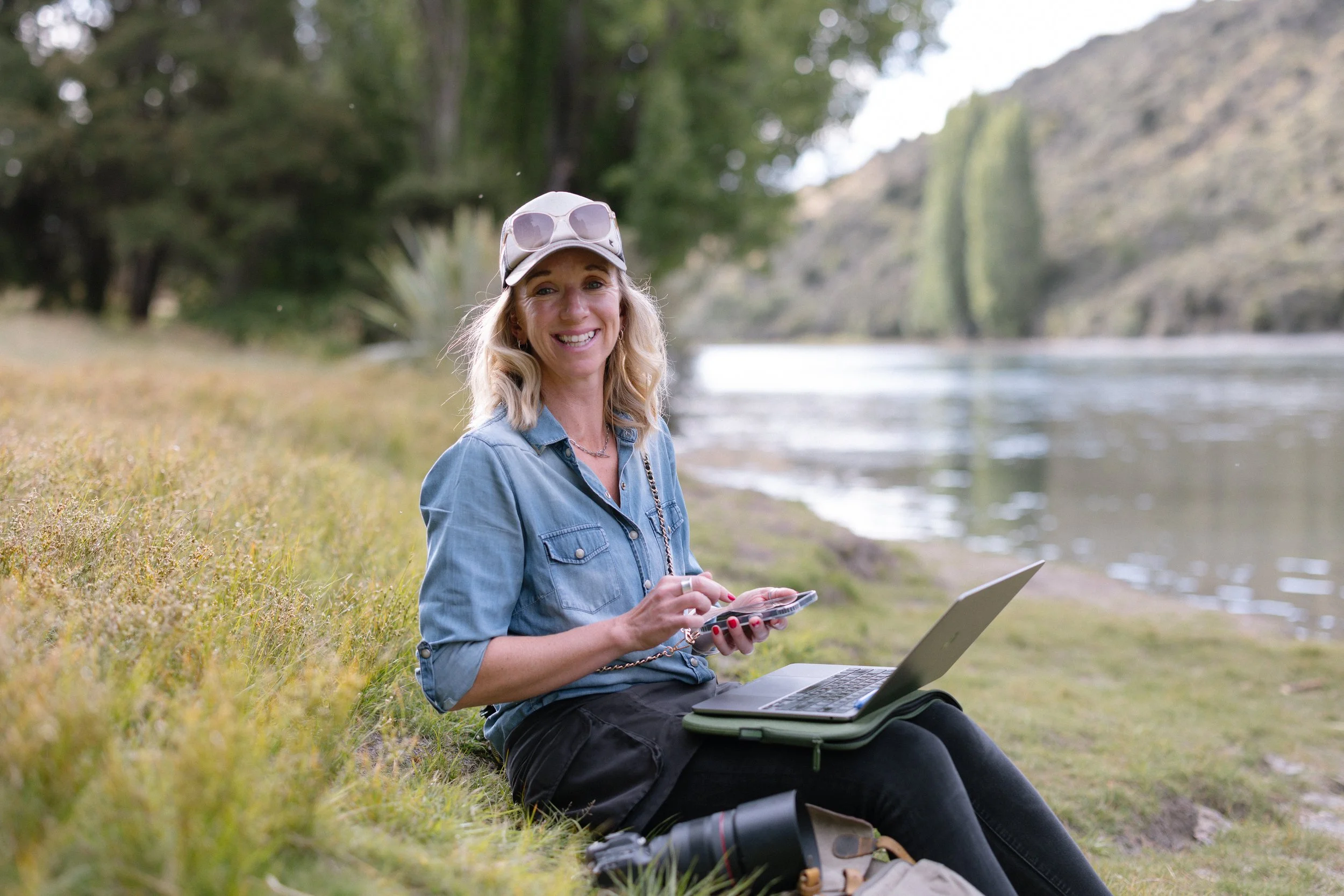 Anna, wearing a gray cap with sunglasses, denim shirt, and black pants, is sitting on the grass near a river, smiling and using her phone, with a laptop on her lap and a camera on the ground nearby in a natural outdoor setting.
