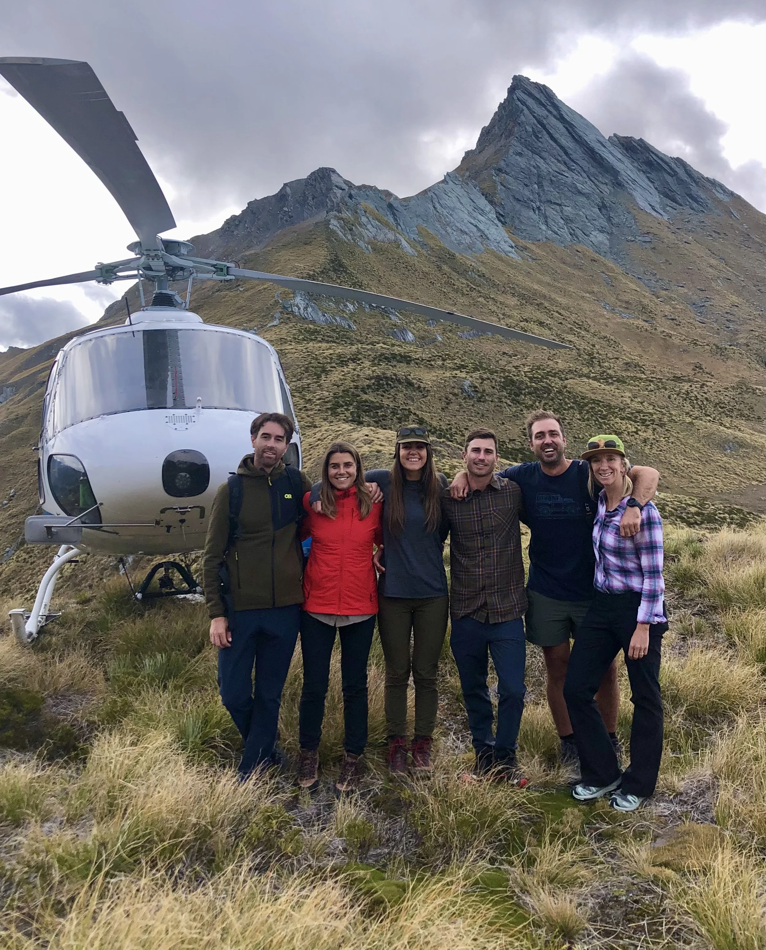 Group of six people smiling and posing in front of a helicopter with mountains in the background, during overcast weather.