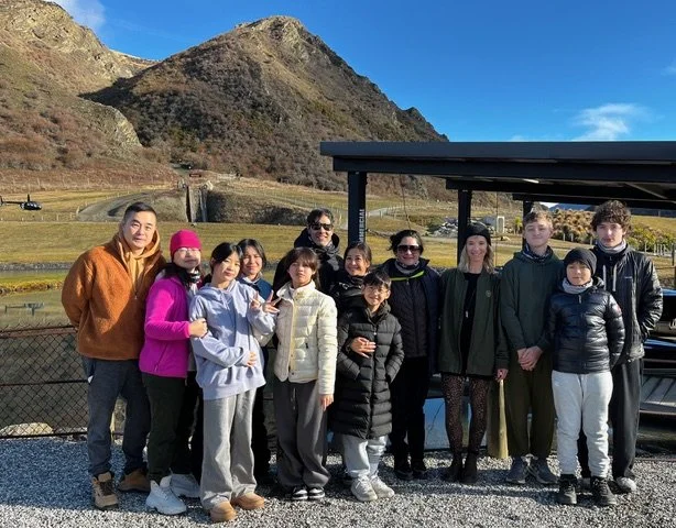 Group of ten people, including children and adults, standing outdoors on a gravel area near a picnic shelter, with a mountainous landscape in the background and blue sky.