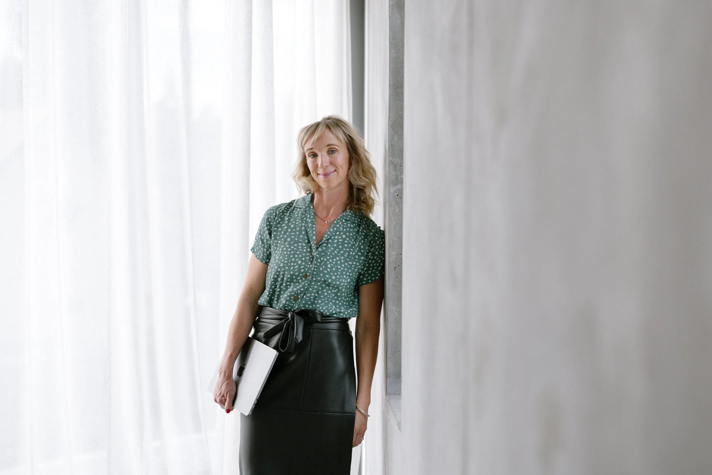 Anna wearing a green polka-dot blouse and black skirt, holding a notebook, standing near a wall with a light-colored curtain in the background.