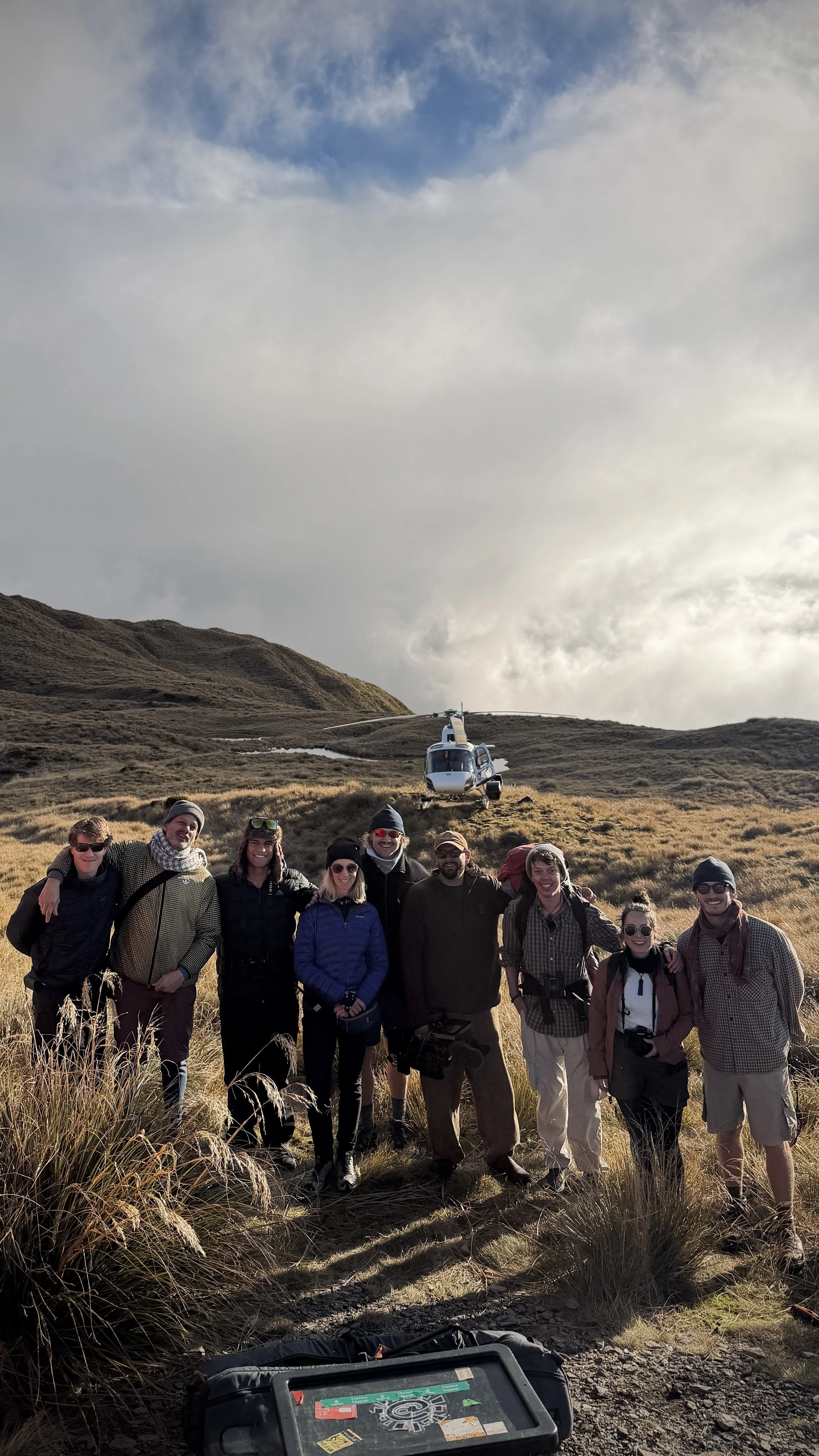 Group of people standing on a hillside with a helicopter hovering behind them and rugged terrain in the background.