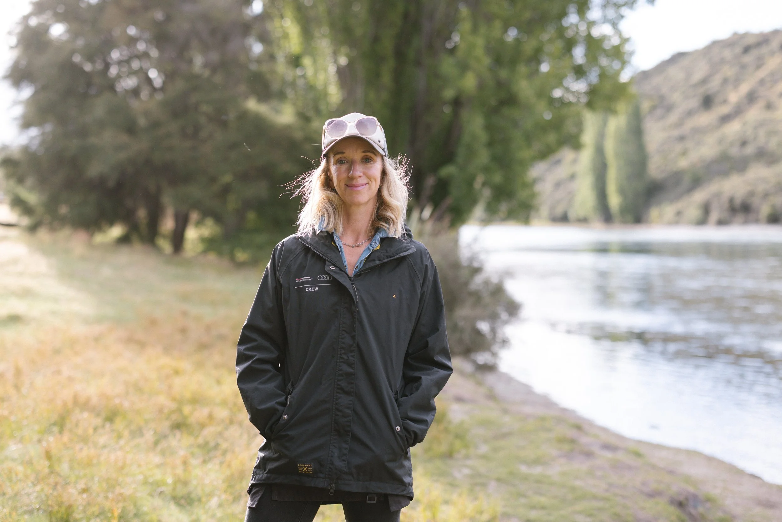 Anna standing outdoors near a river, wearing a black jacket and sunglasses on her cap, with trees and hills in the background.