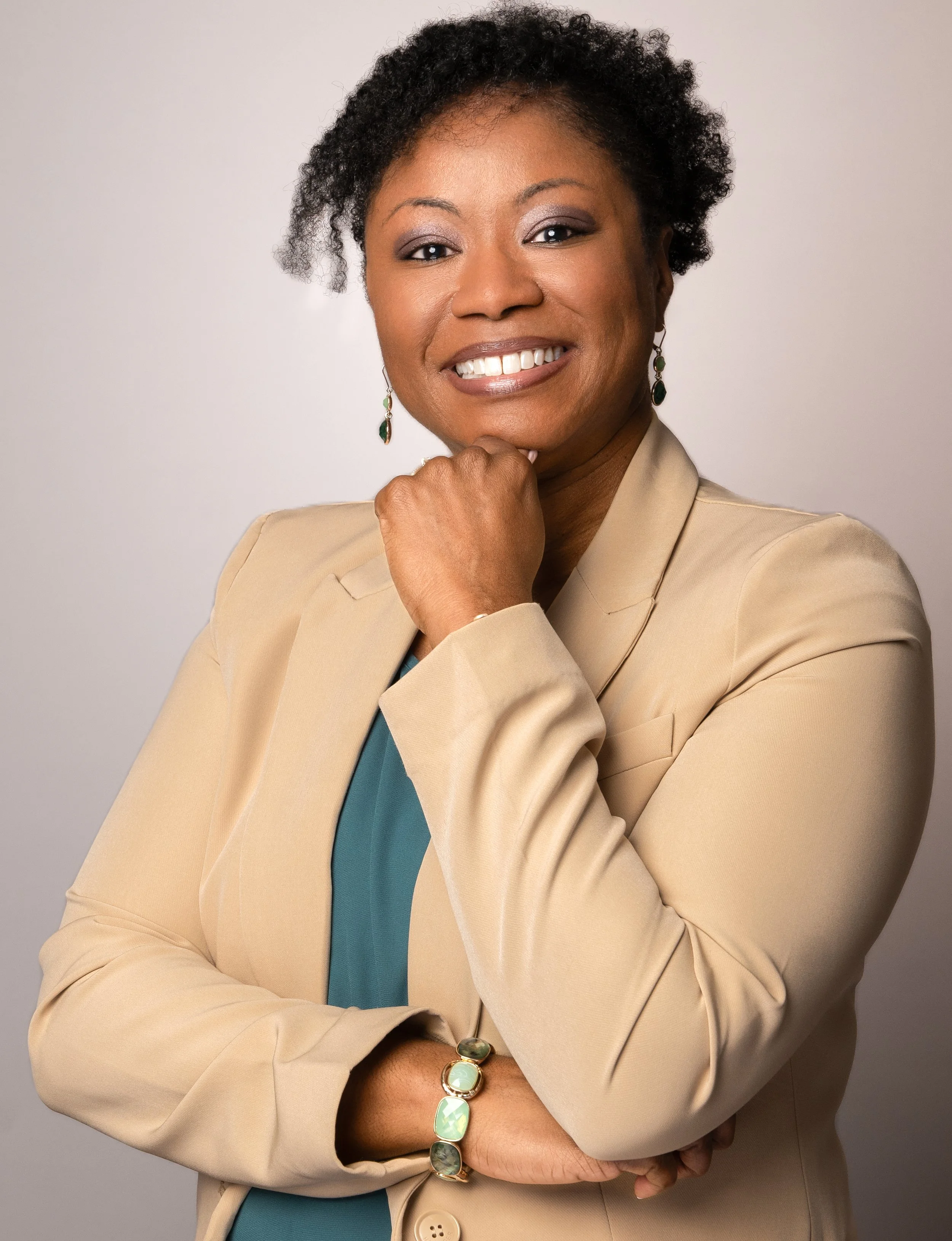 Dr. Lisa Petit, M.D.- Pediatrician and Keynote Speaker in a beige blazer smiling and resting her chin on her hand, wearing earrings and a bracelet, against a plain light background.