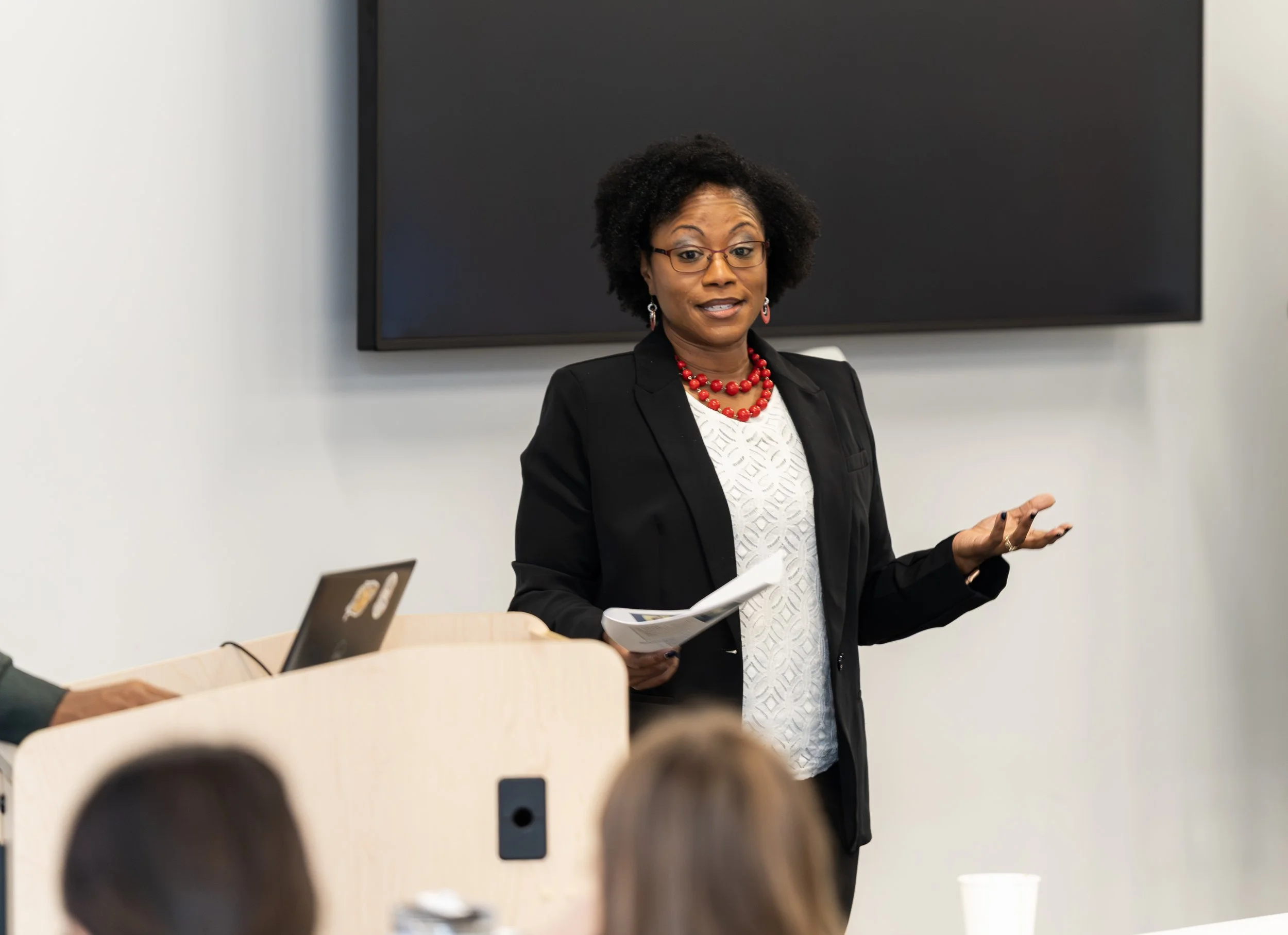 Dr. Lisa Petit, M.D.- Pediatrician and Keynote Speaker in a black blazer and white patterned top stands in front of a blackboard, speaking and gesturing with her hands during a presentation in a classroom or conference room.
