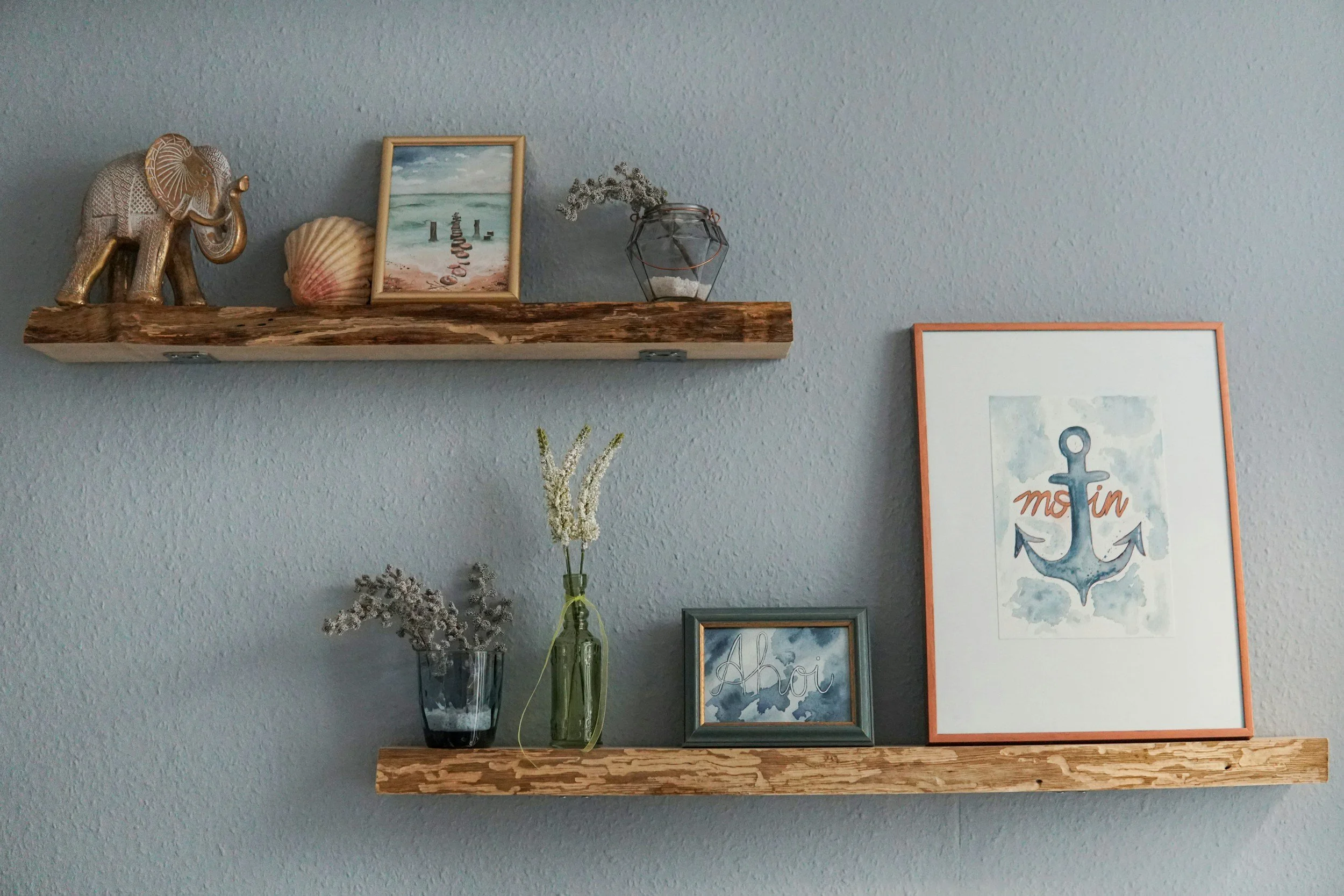 Decorative shelf on a wall with a metallic elephant sculpture, a seashell, a framed painting of the beach, a glass jar with dried flowers, and greeting cards with an anchor and a whale illustration.