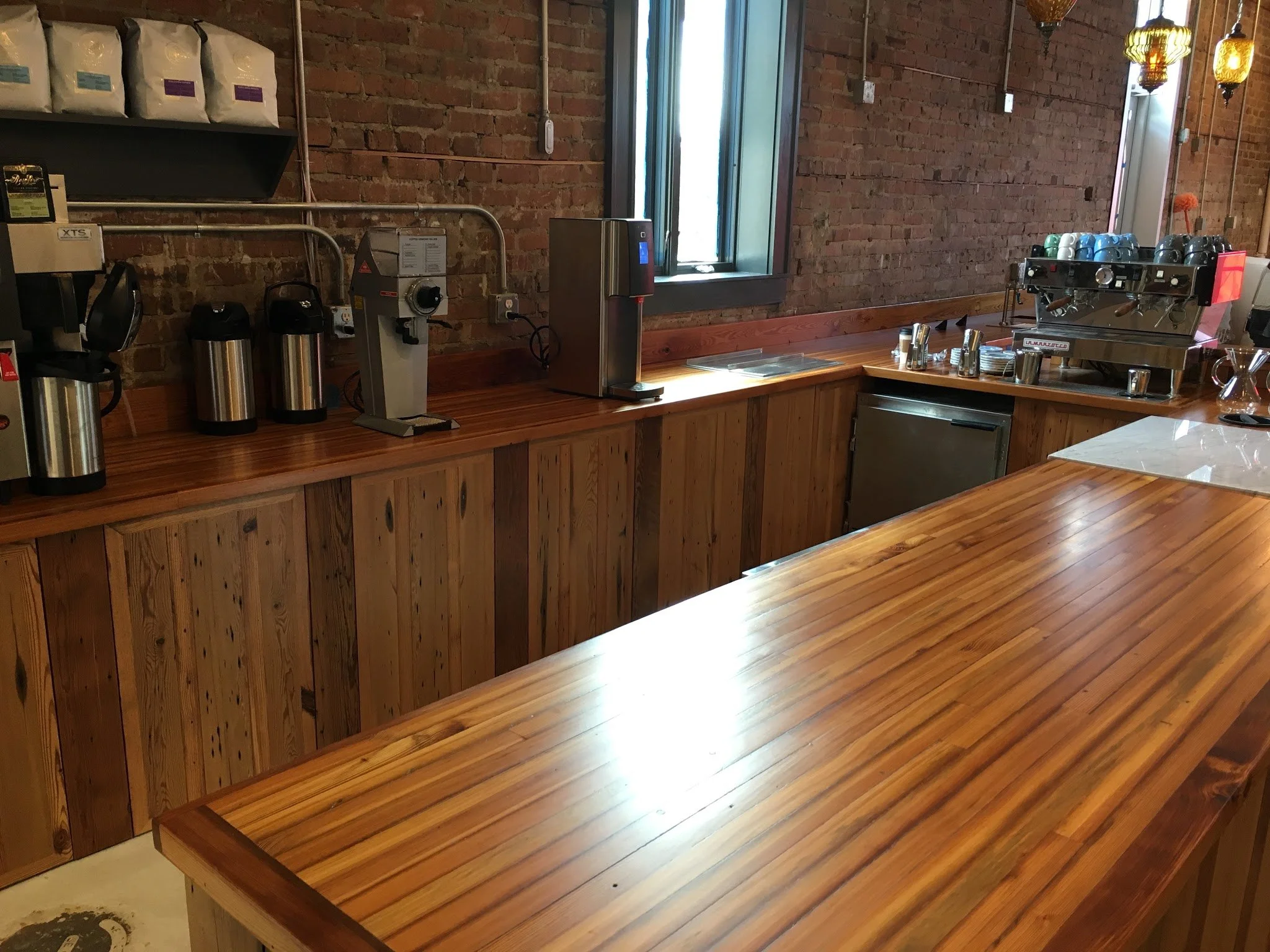 Interior of a coffee shop with a wooden counter, espresso machine, coffee cups, and various coffee-making equipment. Exposed brick walls and nearby window letting in natural light.