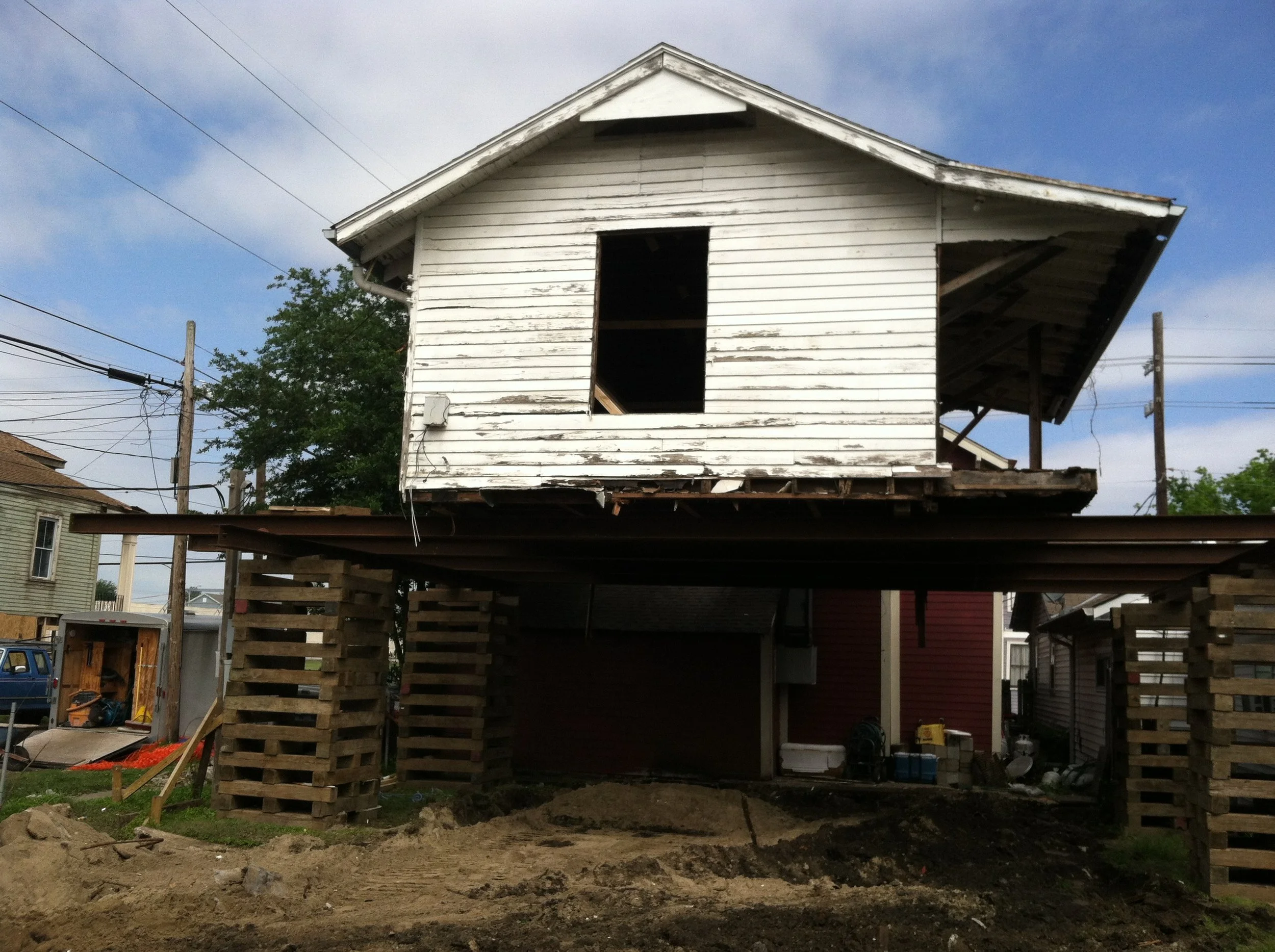 An under-construction house with an elevated foundation made of wooden pallets. The house has weathered white wooden siding and a black rectangular opening where a window or door will be installed. Surrounding the construction site are power lines, t