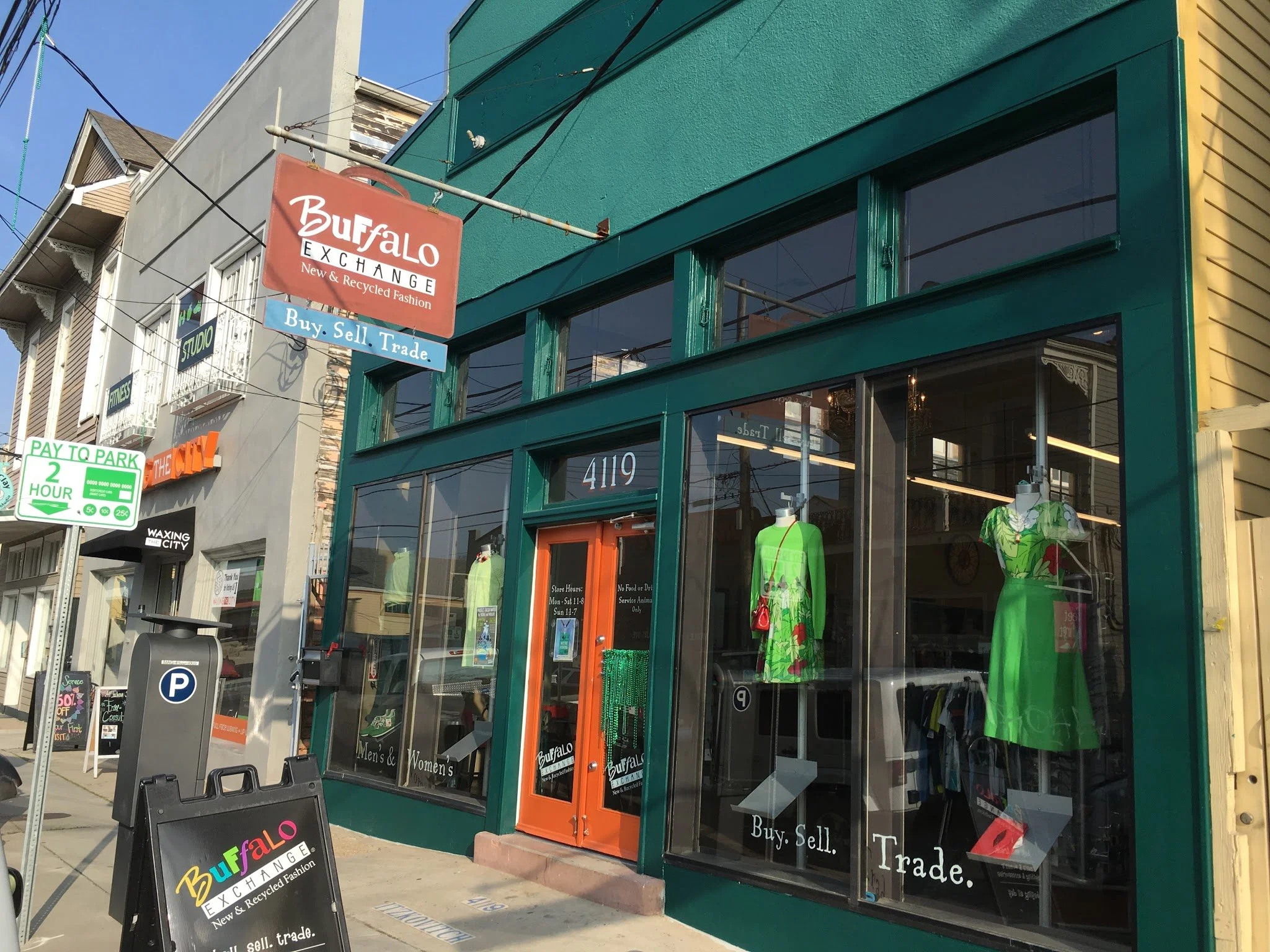 Street view of Buffalo Exchange store with green exterior, large windows, mannequins in colorful clothing, and a red sign that reads "Buffalo Exchange, New & Recycled Fashion" with a smaller blue sign below that says "Buy. Sell. Trade."