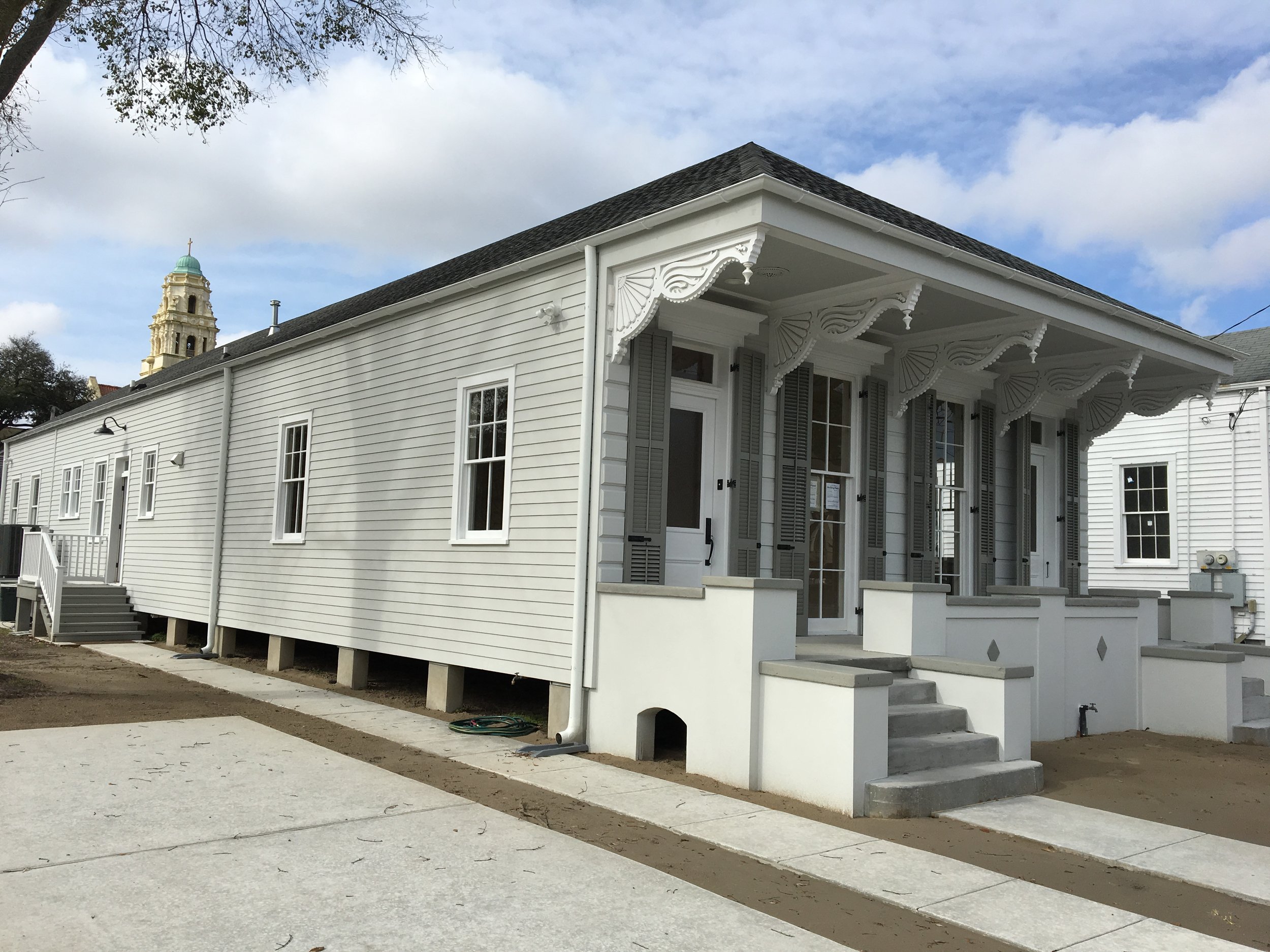 A white house with decorative wooden trim and gray shutters, set against a partly cloudy sky. A church steeple is visible in the background. The house has steps leading to a porch and a concrete walkway in front.