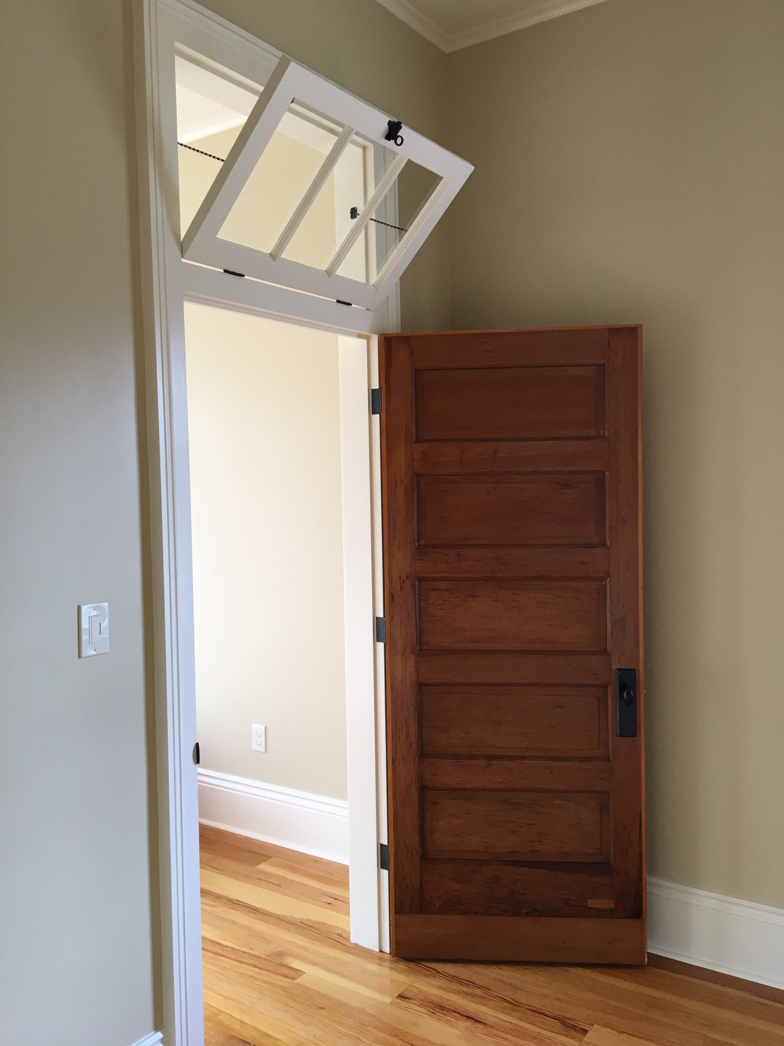 Open wooden door in a room with light-colored walls and hardwood floors, showing an adjoining room with a similar light-colored wall and hardwood flooring.