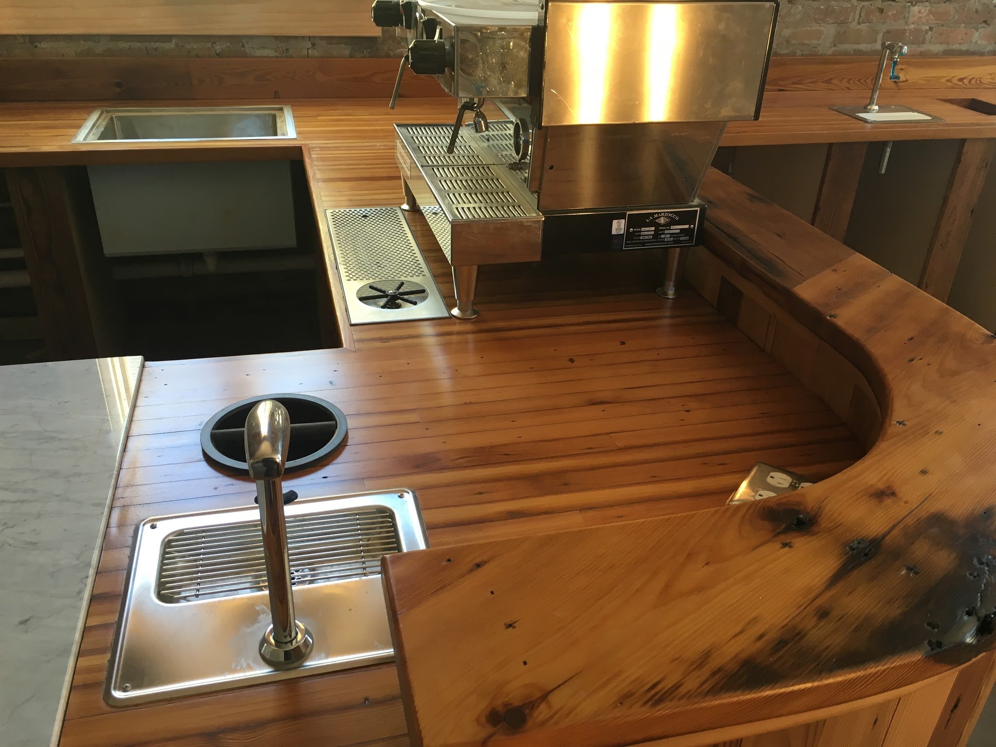A wooden bar counter with a stainless steel espresso machine and a matching drip tray. There is a circular drain cover on the counter, a small sink to the right, and an electrical outlet with power cords at the bottom.