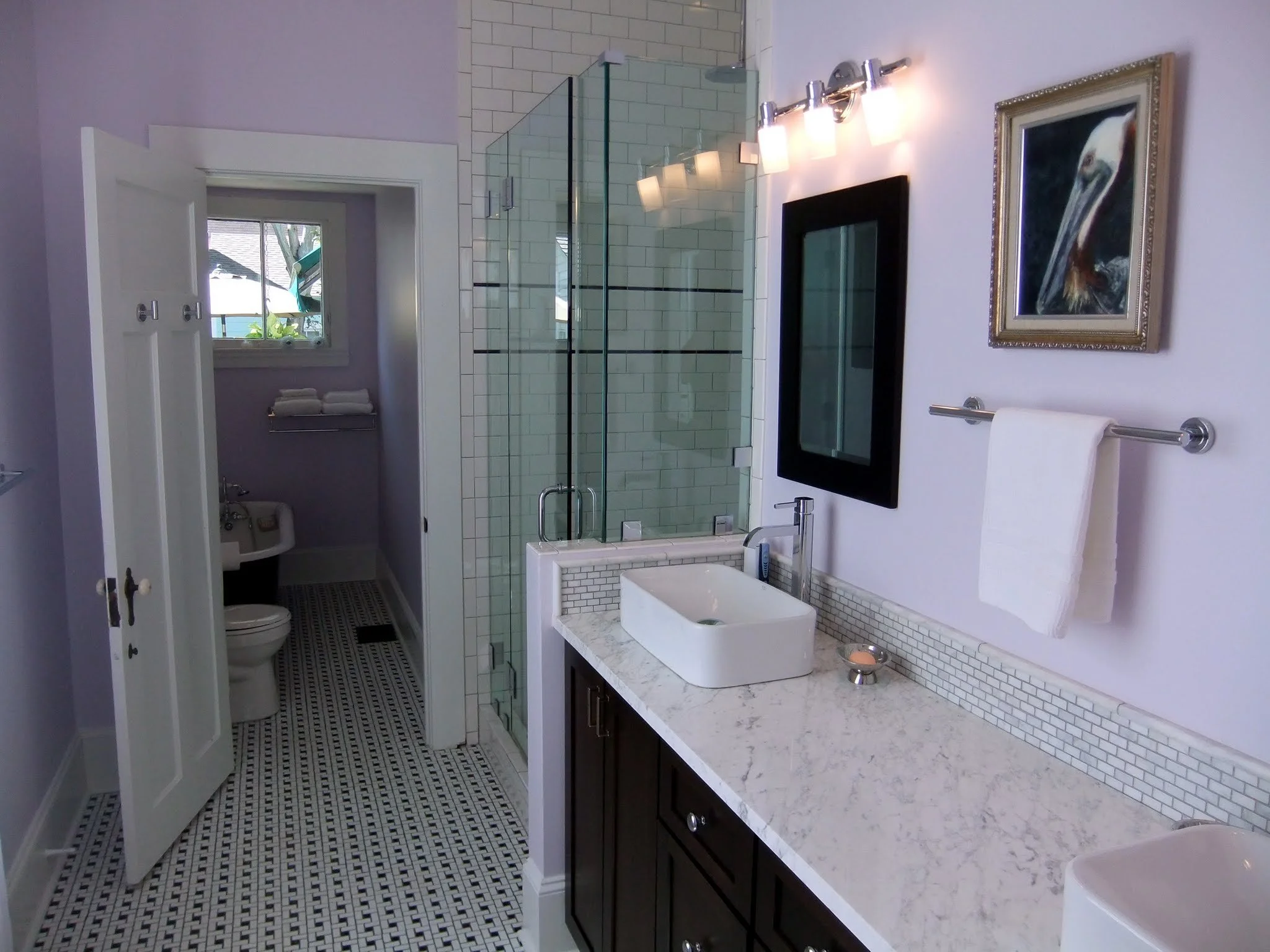 Bathroom with a marble countertop sink, dark wooden cabinets, a large mirror, white tiled shower with a glass enclosure, a window, a white towel on a towel bar, a framed picture of a heron, a wall-mounted light fixture, and a soap dish with soap on t