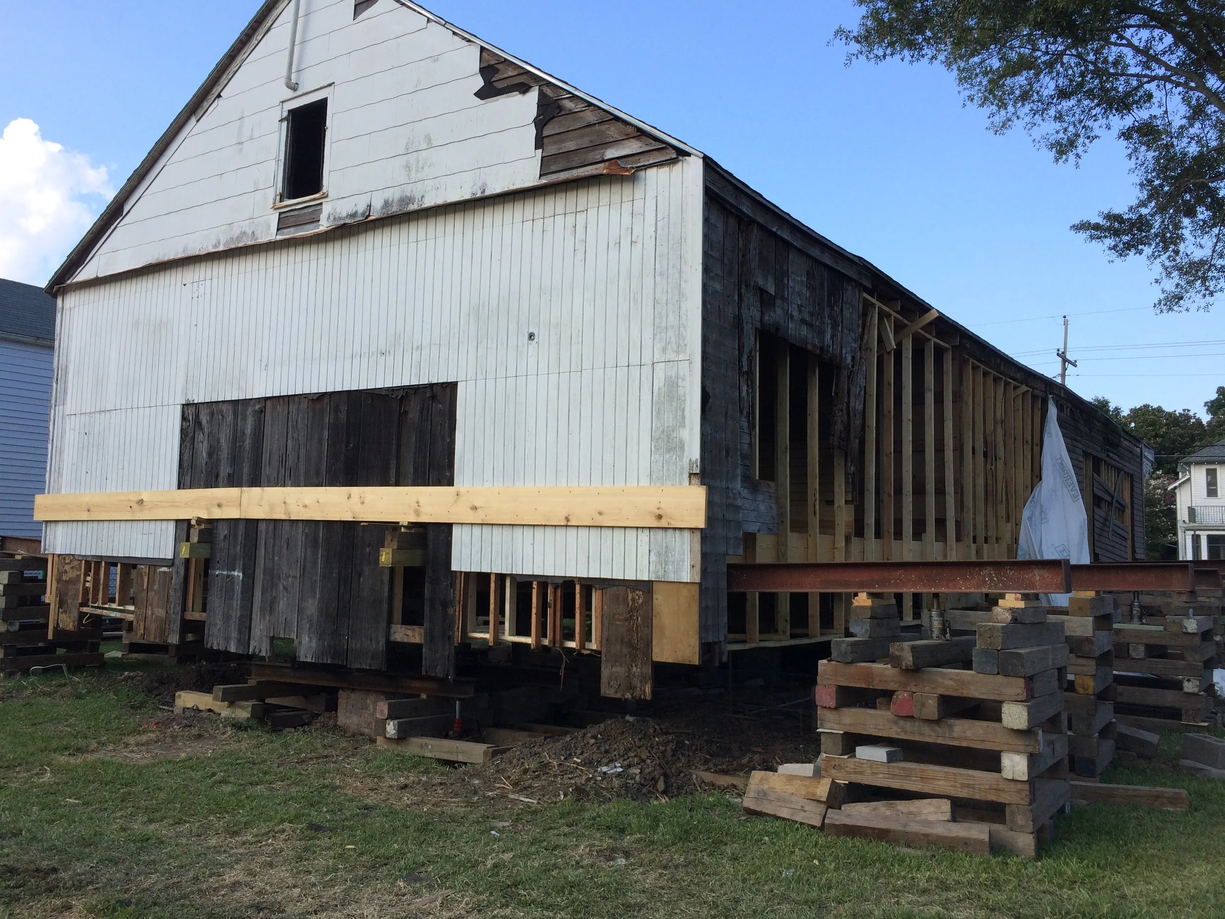 A building under construction, with some parts covered in white siding and some parts in dark weathered wood. The structure is raised on wooden and concrete supports, and the roof is in the process of being finished. The sky is clear with a few cloud
