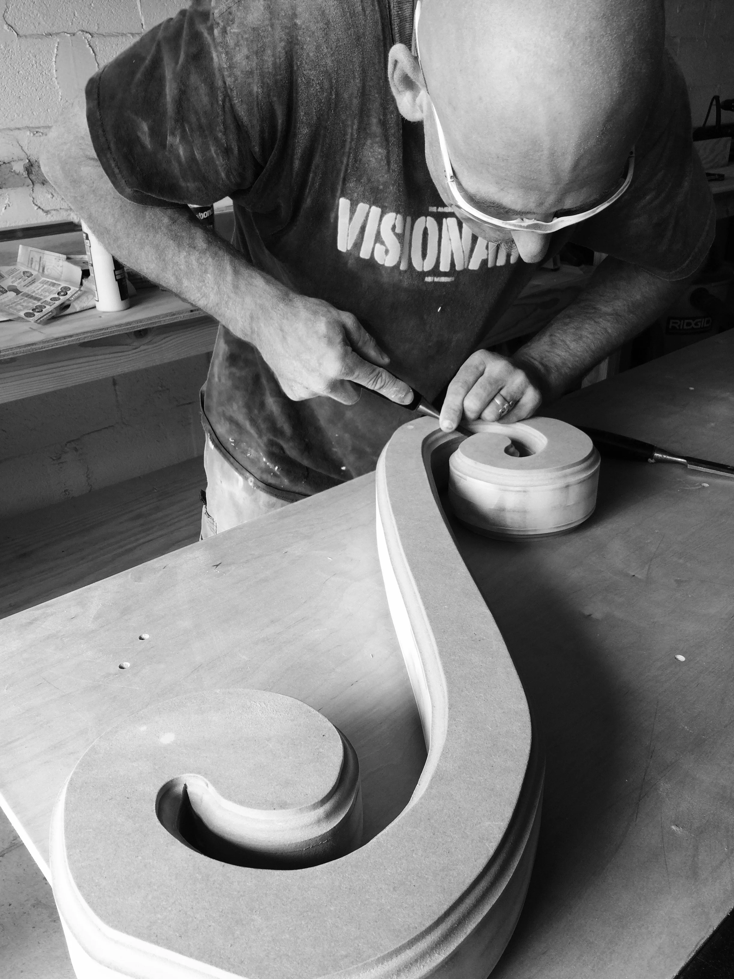 Man wearing glasses and a logo t-shirt carving into a piece of wood with a saw in a woodworking workshop.