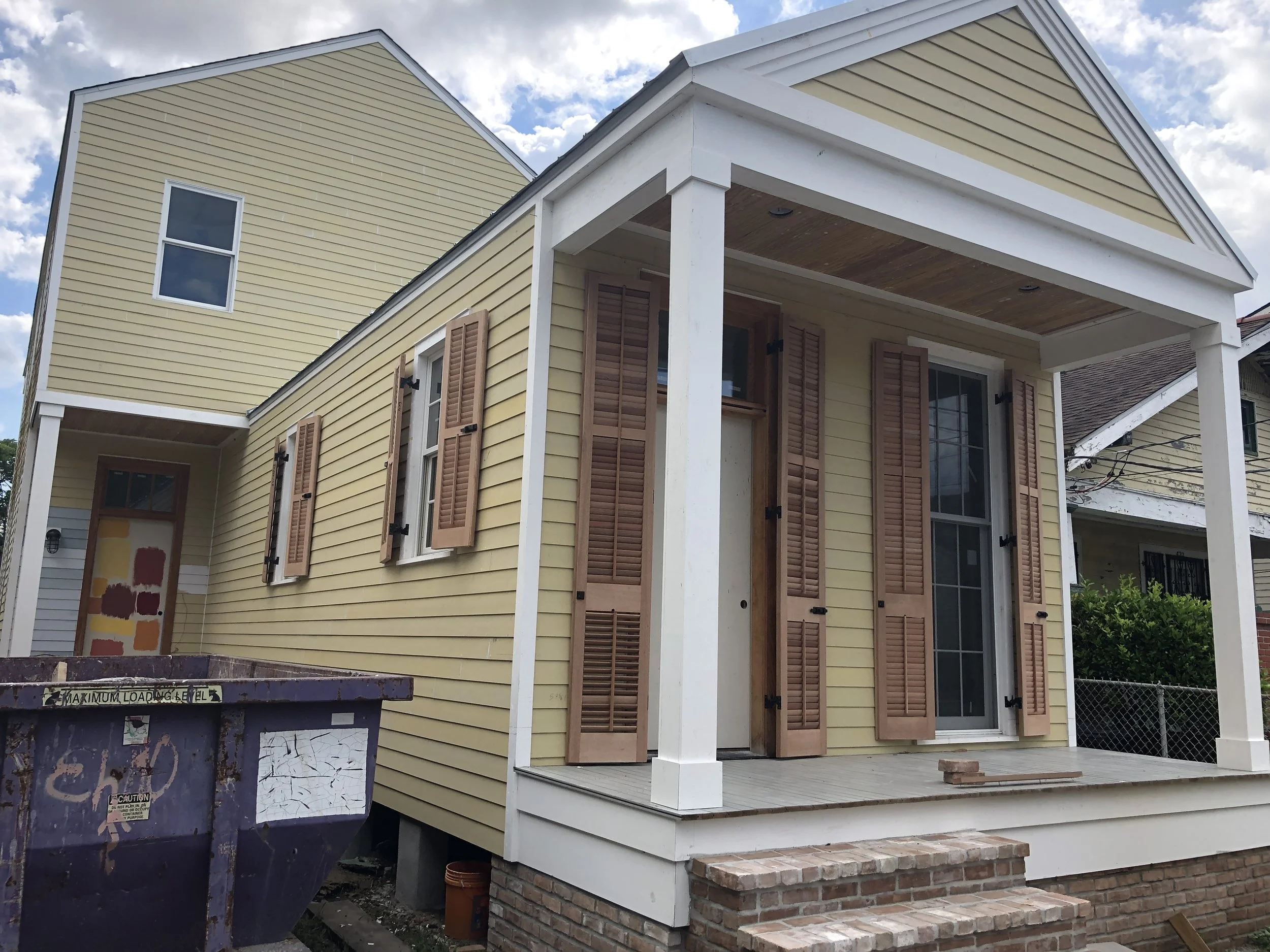 The image shows a two-story house with yellow siding, brown shutters on the windows, a covered front porch with white columns, and a brick foundation. There is a blue dumpster in the foreground and a cloudy sky overhead.