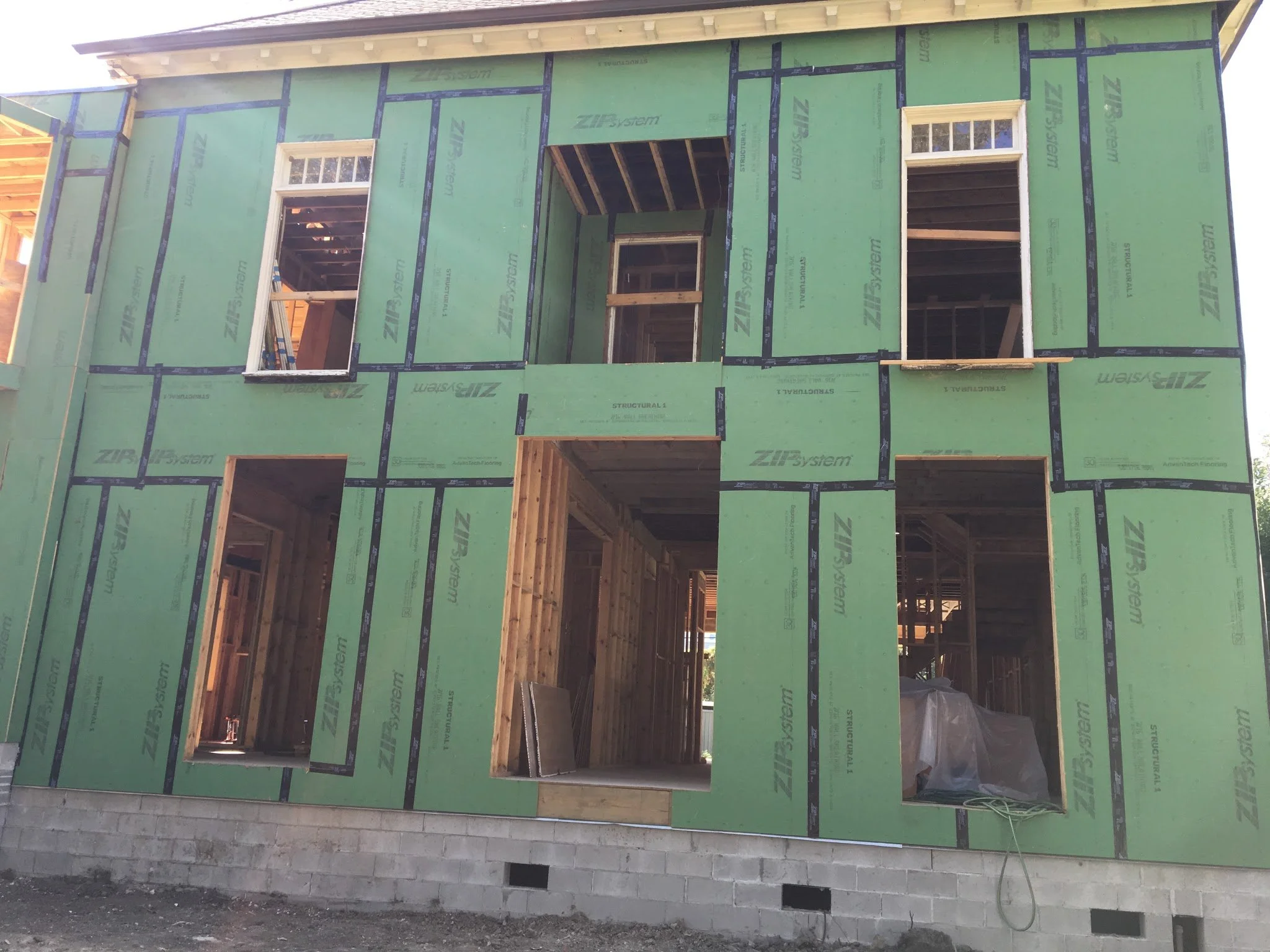 Front view of a house under construction with green sheathing panels, multiple window openings, and a partially completed doorway.