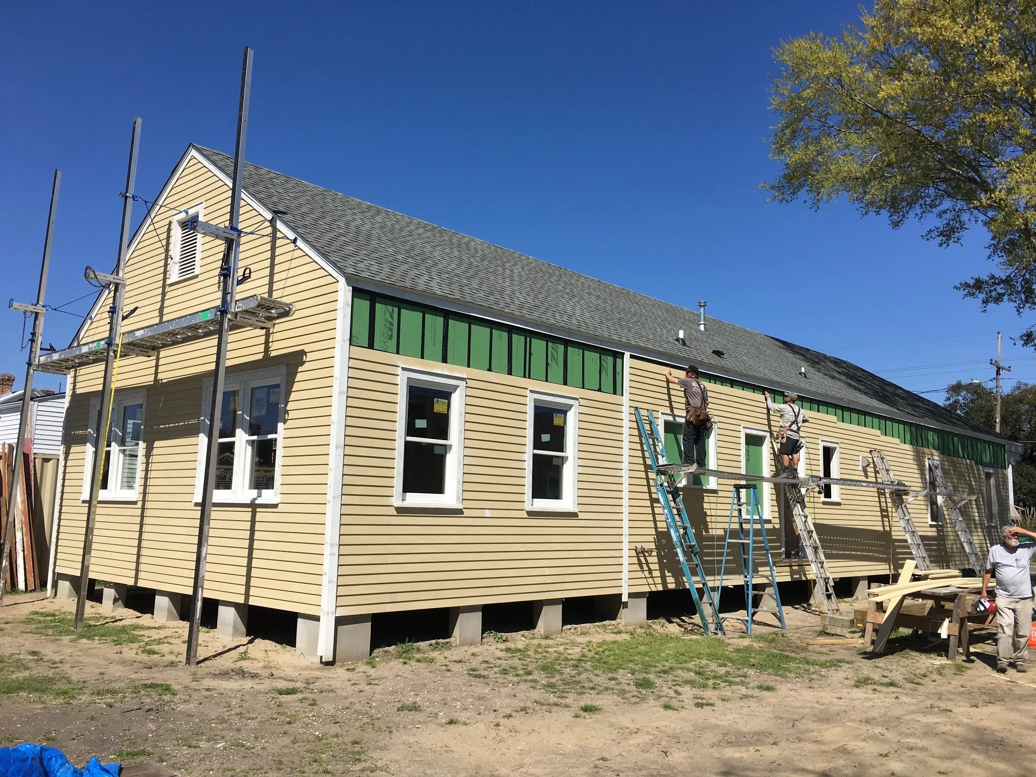A house under construction with workers installing windows on the upper floor, scaffolding set up around the building, and a ladder leaning against the wall. The house is yellow with white trim and has a gabled roof. There are trees and a clear blue 