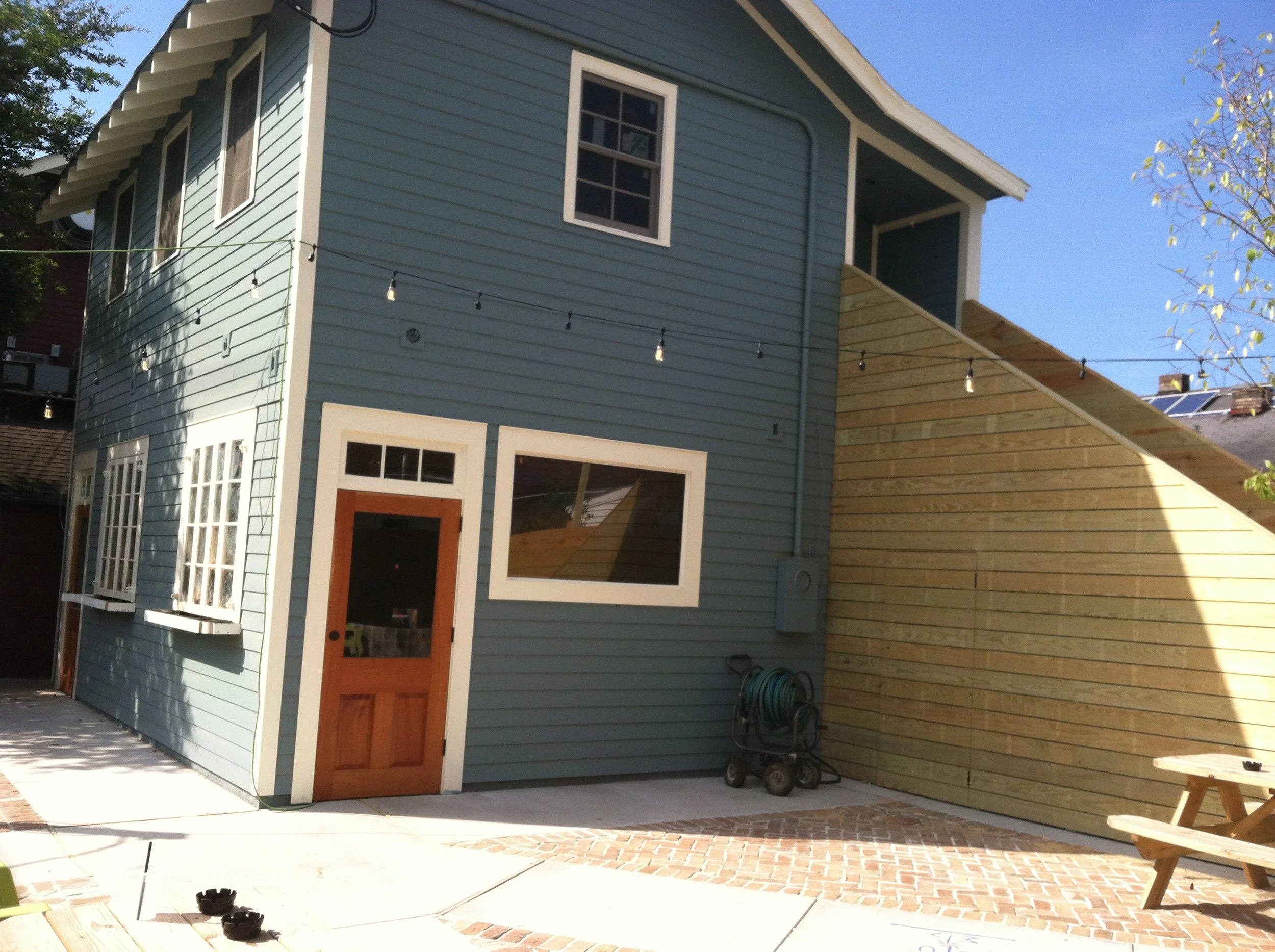 A two-story house painted light blue with white trim. There are multiple windows, a wooden front door, and a small covered balcony on the second floor. Outside, there are string lights hanging, a garden hose, and some outdoor furniture and tools. A s