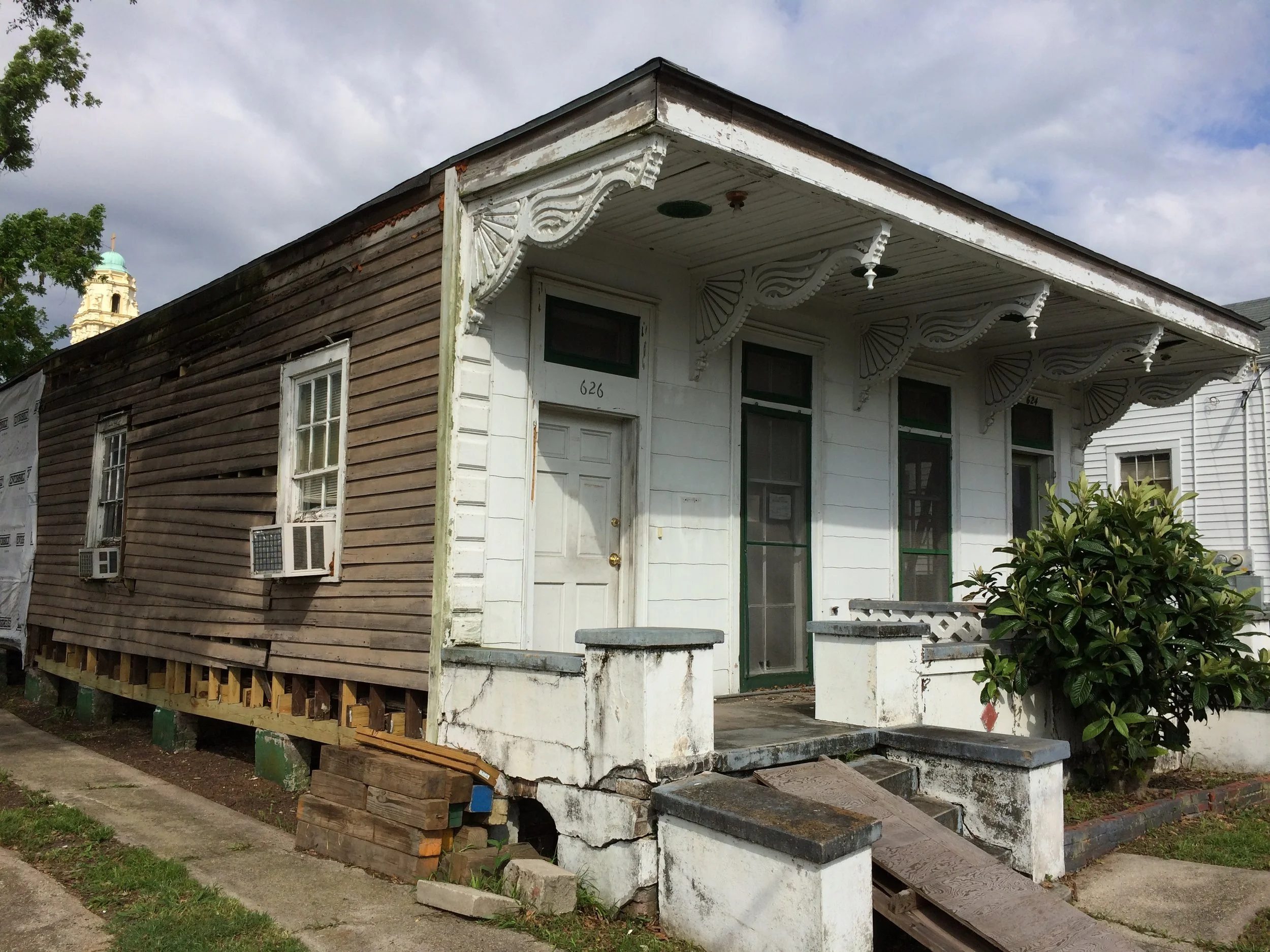An old house with decorative corbels under the eaves, a front porch with stairs, and two air conditioning units in the windows, with some parts of the house under renovation showing exposed wood and foundation.