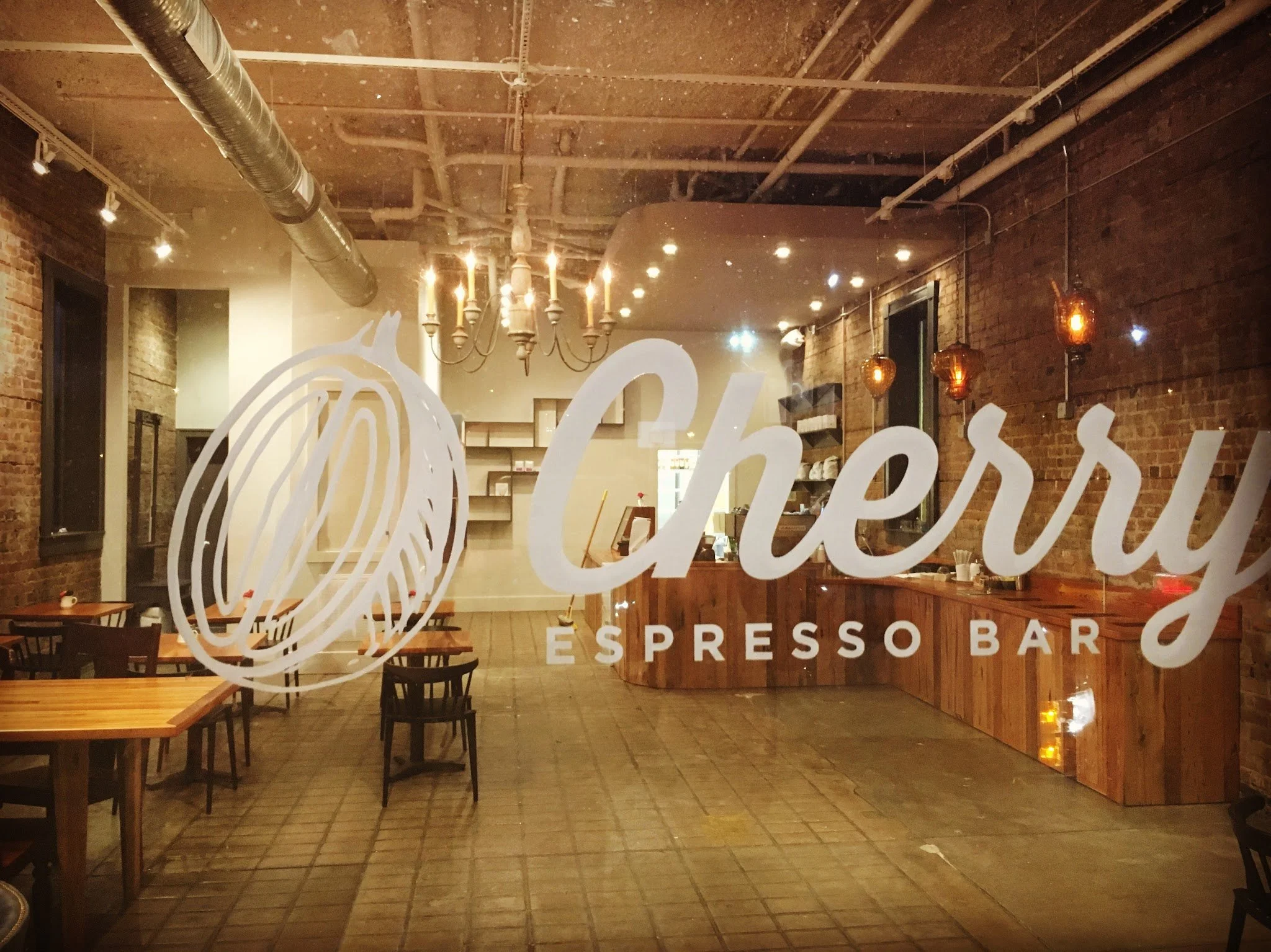Interior of a coffee shop or cafe with brick walls, wooden tables and chairs, hanging pendant lights, a chandelier, and visible industrial ceiling pipes, seen through a window with the text 'Espresso Bar' on it.
