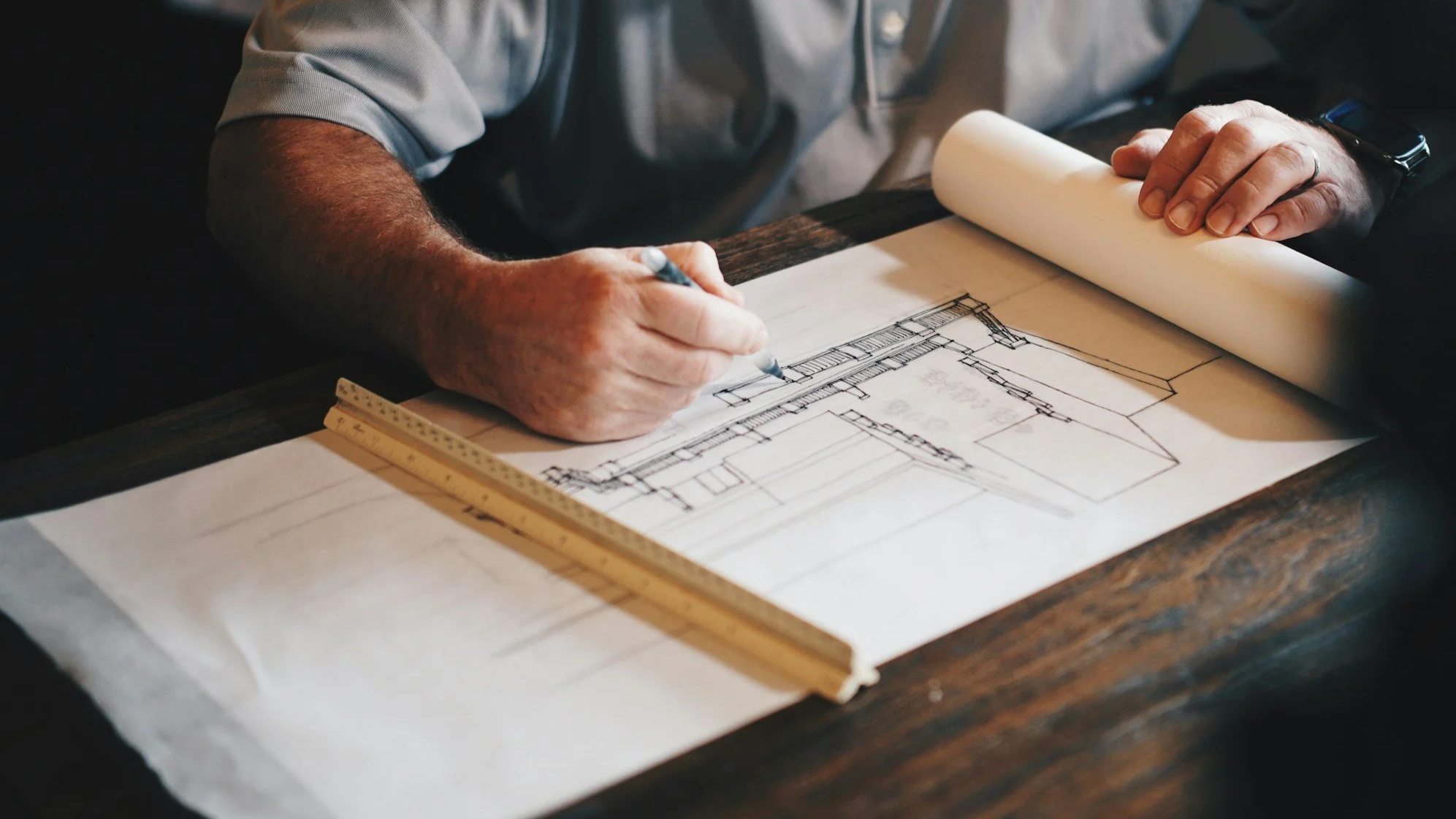 A person drawing architectural blueprints on a piece of paper, with a ruler placed nearby on a wooden table.