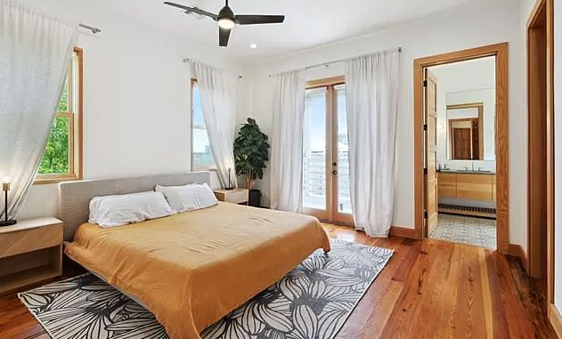 A bedroom with a large bed with white pillows and an orange bedspread, two nightstands, a potted plant, hardwood floors, two windows, and glass doors leading to an outdoor area. There is an adjoining bathroom visible through a door.