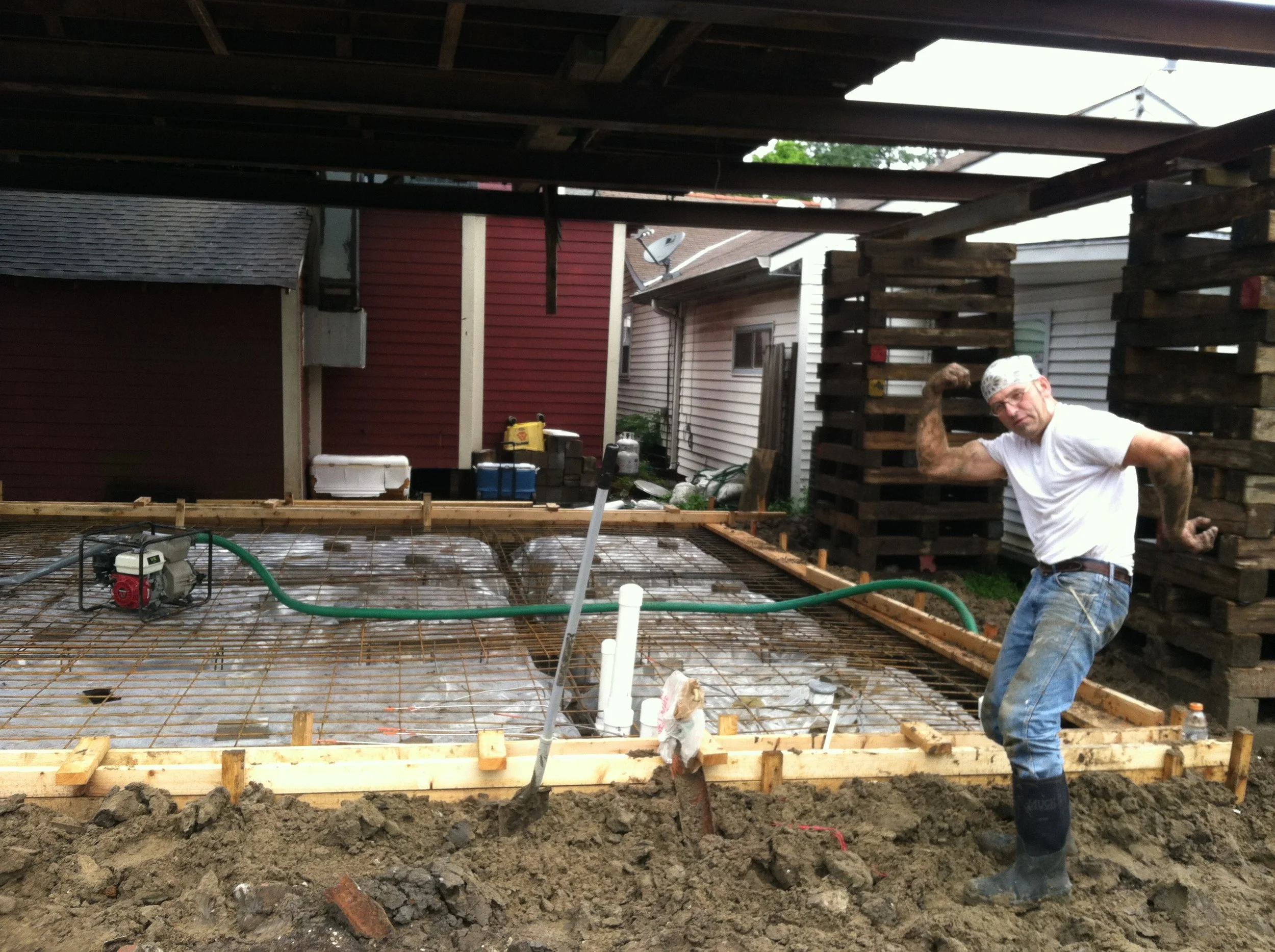 Man flexing his arm muscles while standing on a construction site in front of a foundation with rebar, with houses and trees in the background.