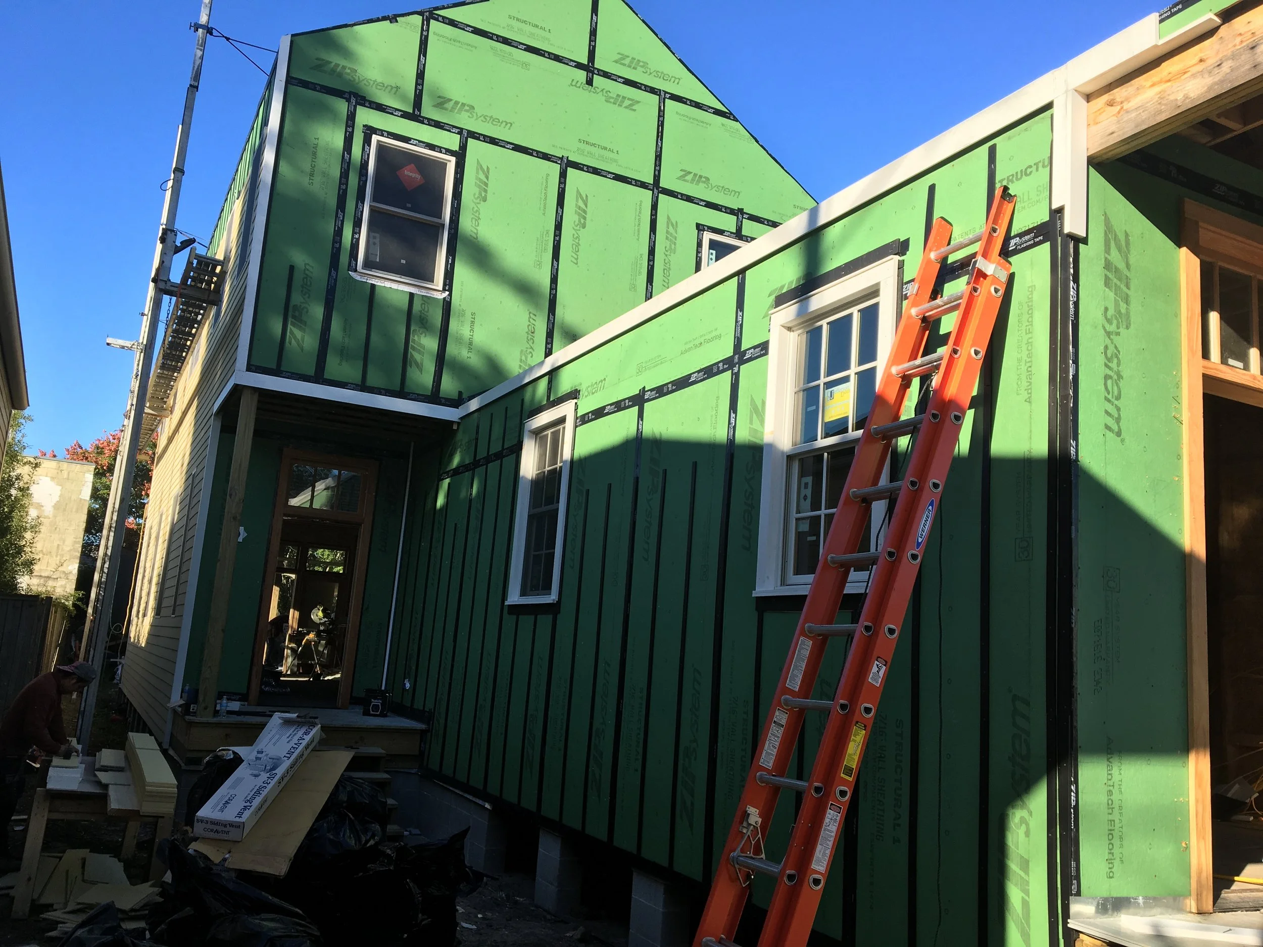 House under construction with green exterior sheathing, an orange ladder leaning against it, and construction workers working nearby.