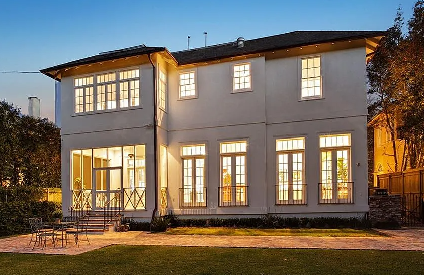 Two-story house with large windows illuminated from inside, a patio with outdoor furniture, and a well-maintained lawn at twilight.