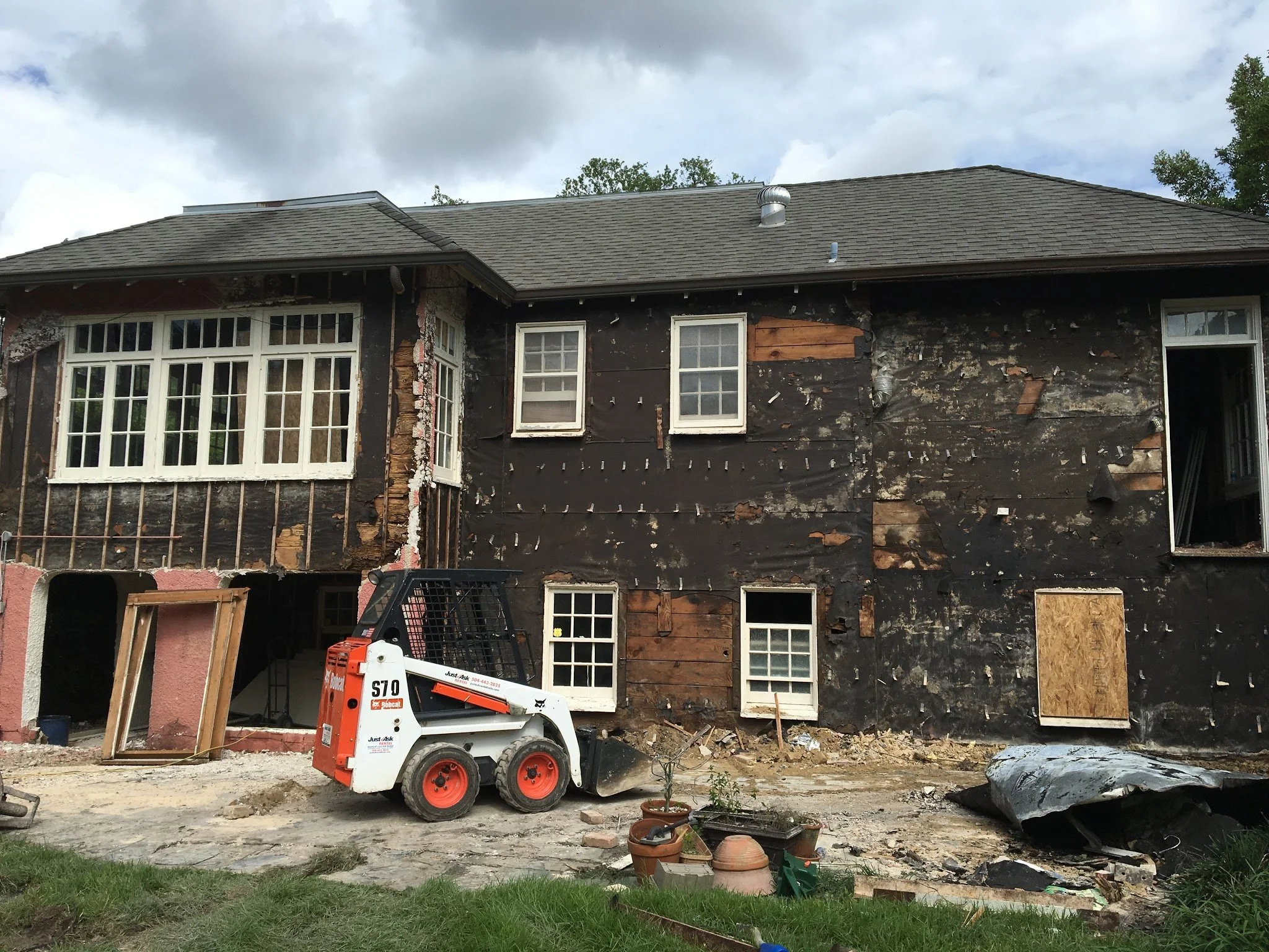 House undergoing renovation with exposed black sheathing, missing siding, and construction equipment such as a Bobcat mini loader in the yard.