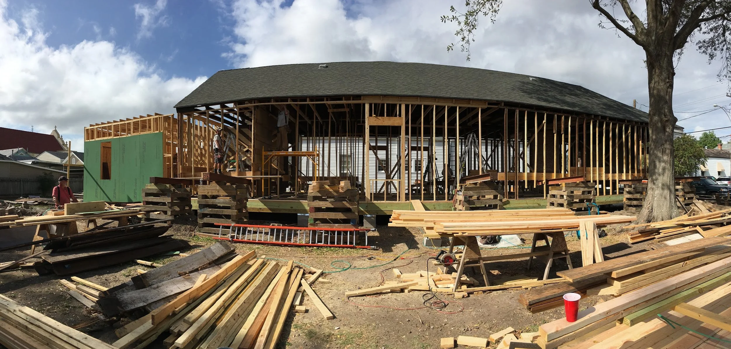 Construction site of a house with wooden framing and workers installing structural elements, surrounded by construction materials and equipment.