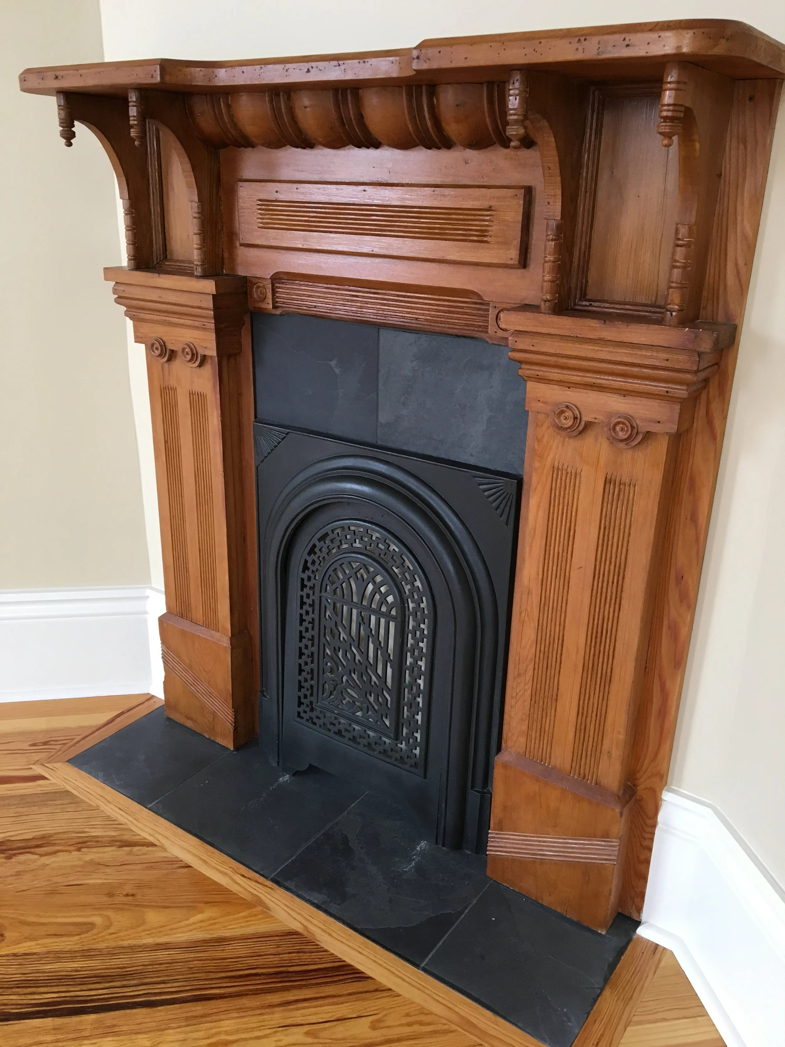 A wooden fireplace mantel with decorative columns and a black fireplace insert, set against a beige wall with a white baseboard and hardwood flooring.