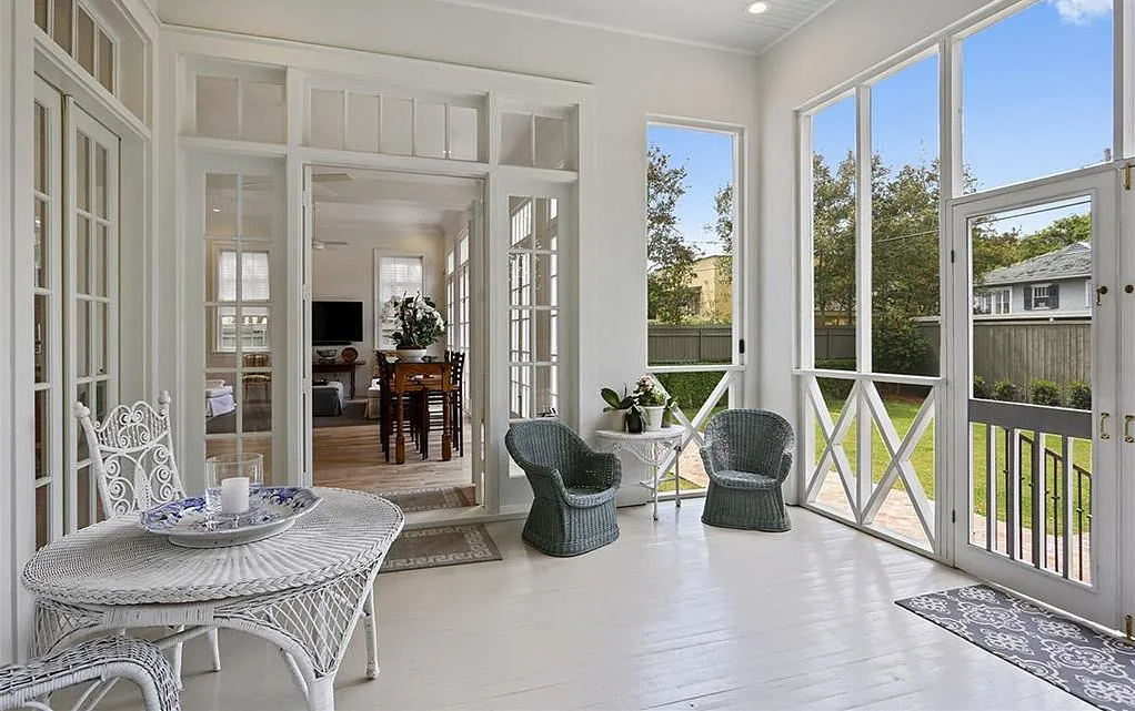 Sunroom with white walls and flooring, enclosed with large windows and a door to the outside. Features wicker chairs, potted plants, a white table, and a dining table area visible through a glass partition.